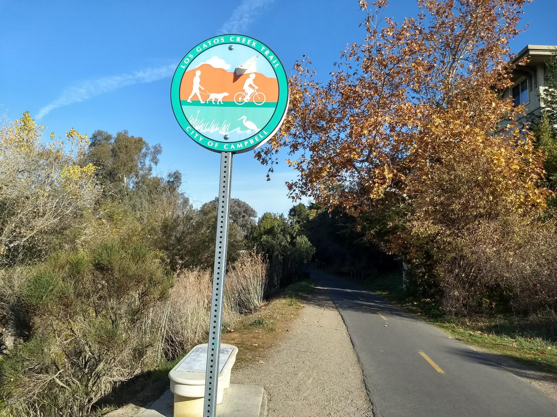 A trail sign stands beside a paved path with autumn trees under blue sky.