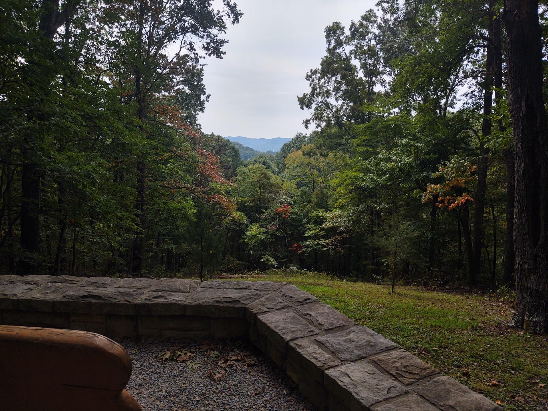 Stone wall overlooking a forest clearing with distant mountains under cloudy sky.