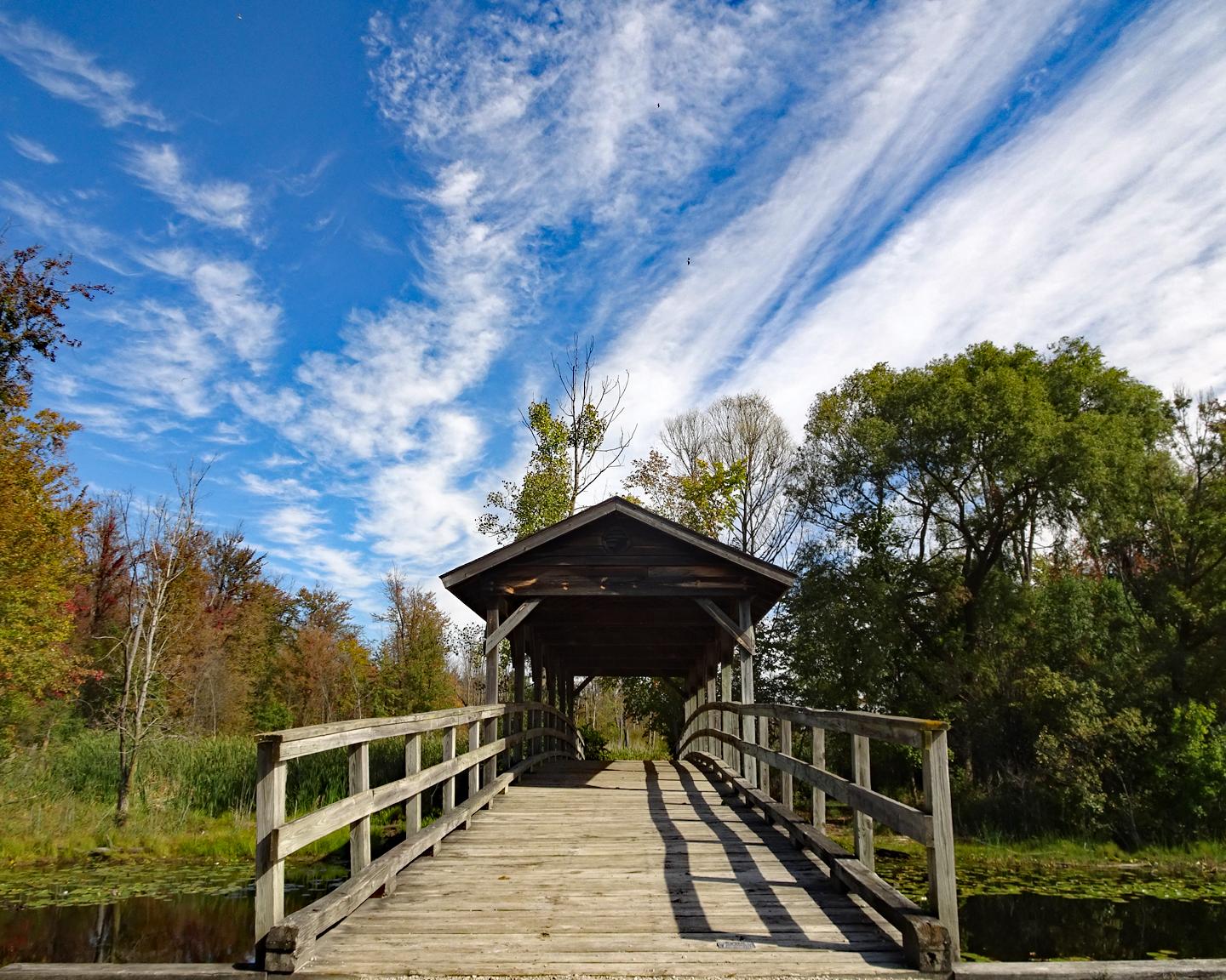 Wooden covered bridge over water, surrounded by trees under a bright, cloudy sky.
