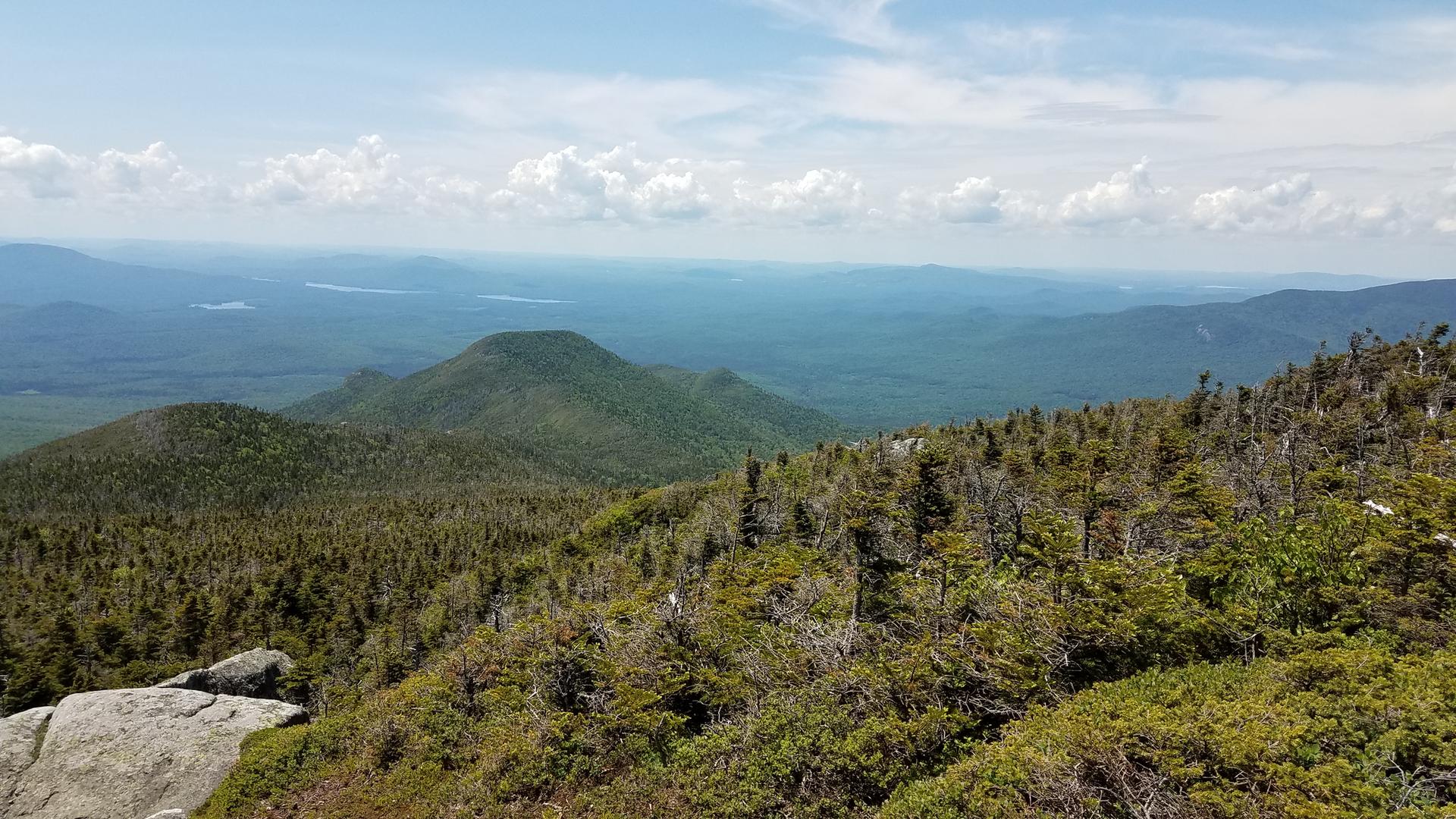 Panoramic view of green forested mountains and hazy distant peaks under a bright sky.
