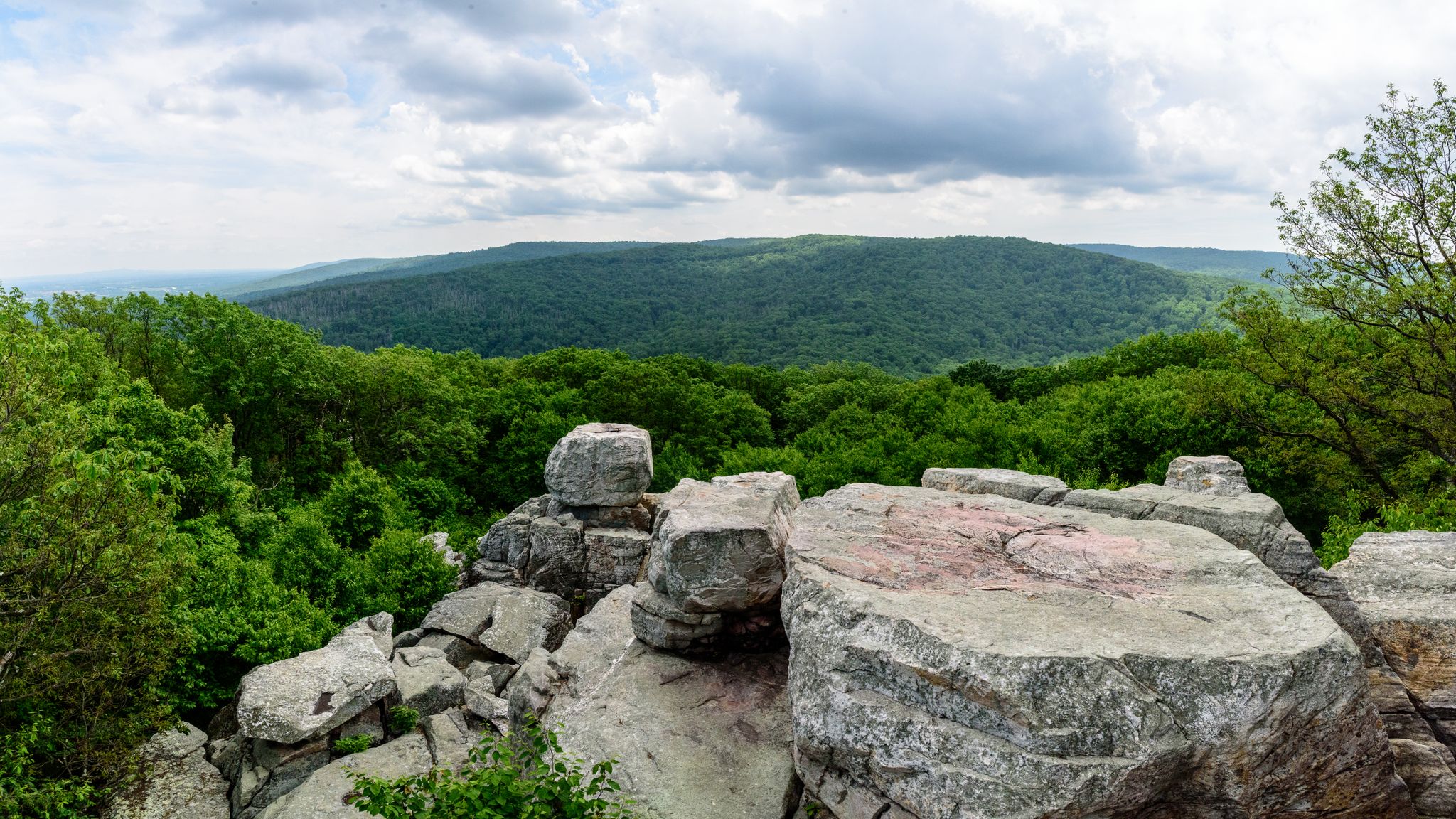 Large rocks overlook a vast, green forested mountain range under cloudy skies. - Orange Trail