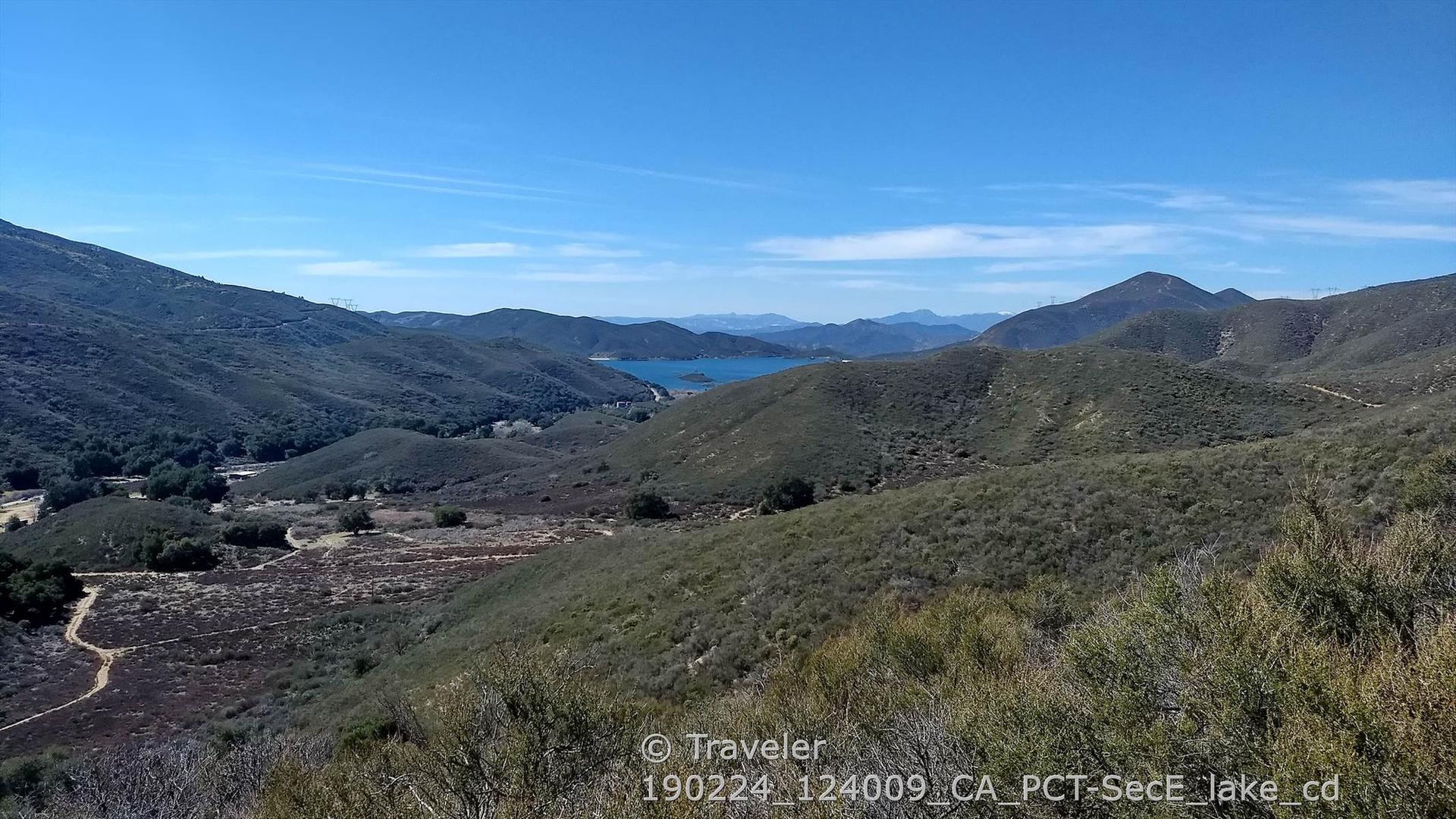 Panoramic view of a large lake nestled between green, rolling mountains under clear sky.