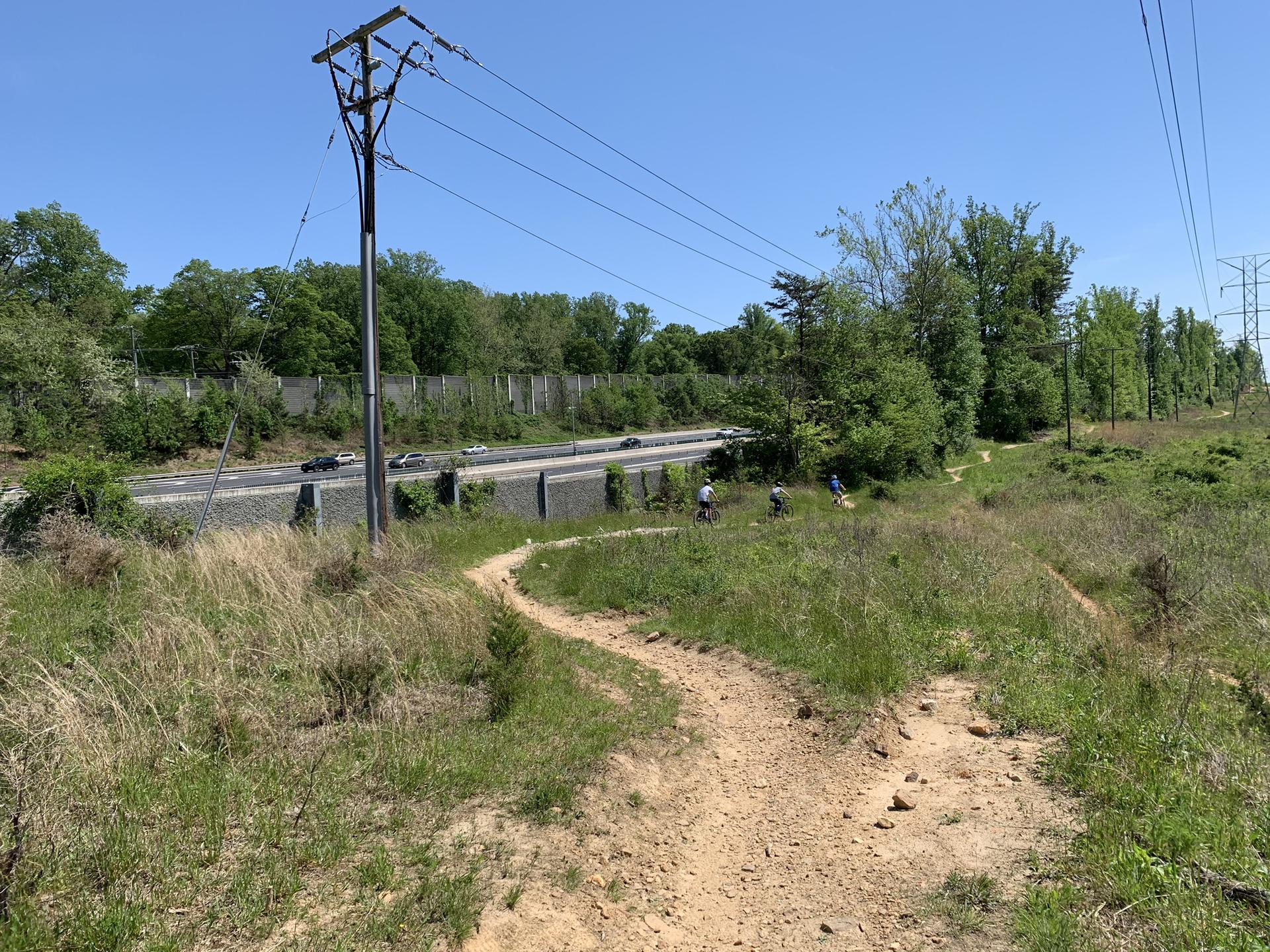 Dirt path with two cyclists, power lines, and a highway on a sunny day.