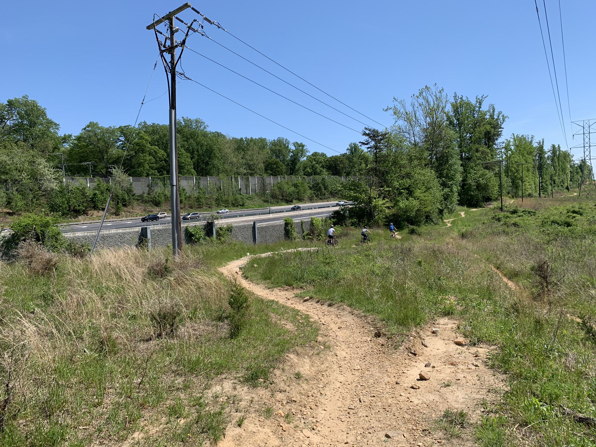 Dirt path with two cyclists, power lines, and a highway on a sunny day. - Wakefield Park MTB Trail