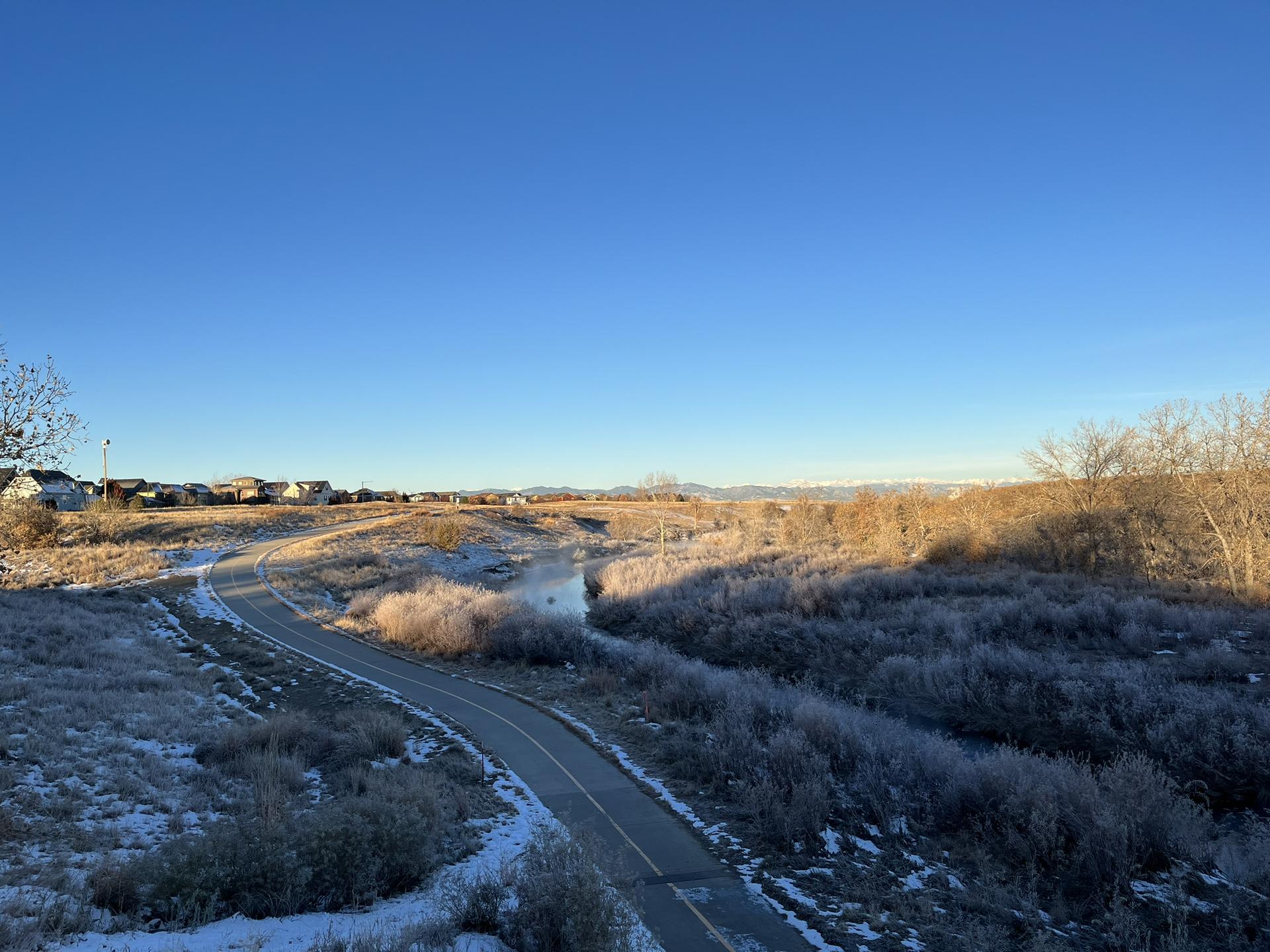 Frosty landscape with a paved path and winding stream under a clear blue sky.