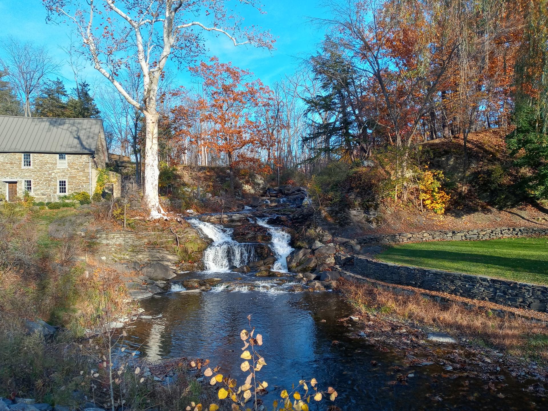 Waterfall cascading past an old stone building and autumn trees.