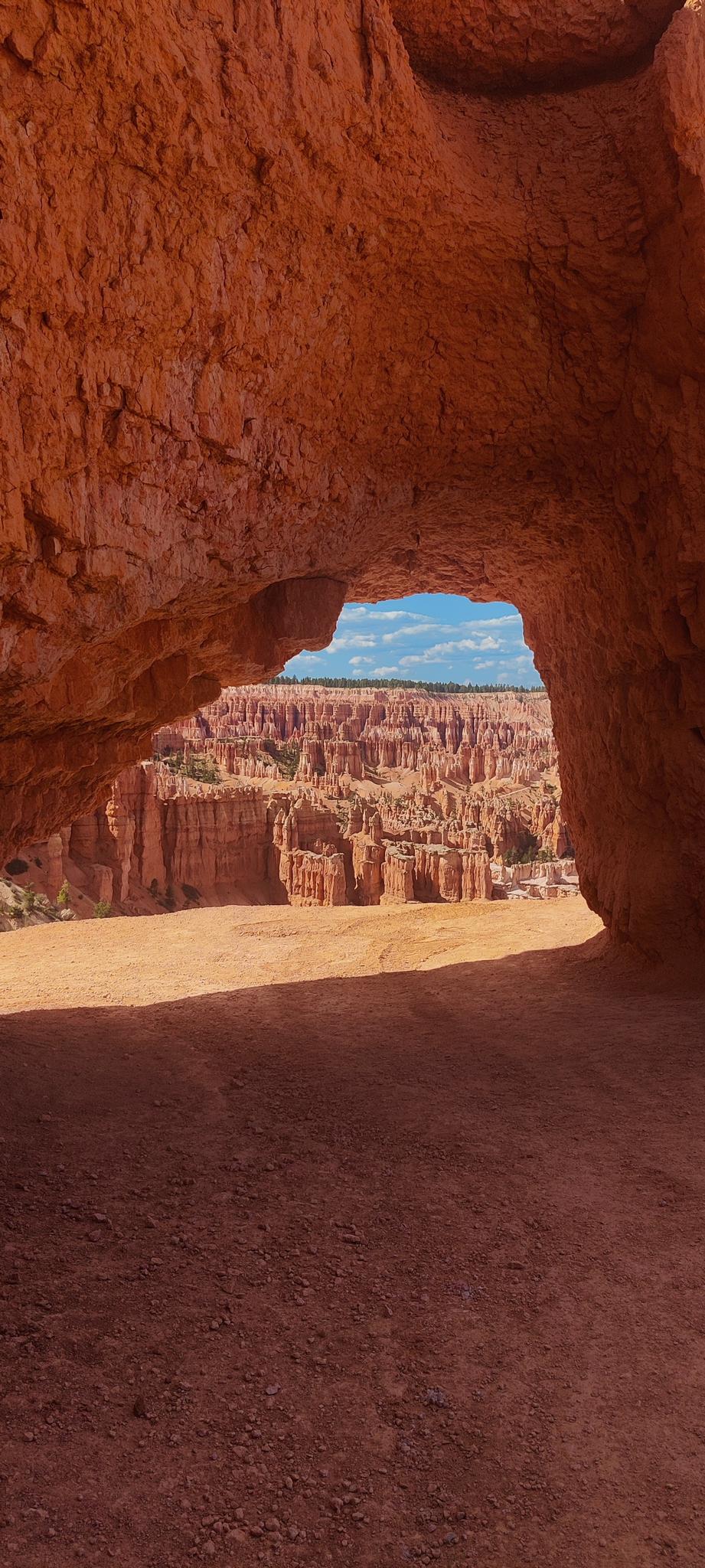 View through a dark red rock arch to sunlit canyon formations and blue sky.