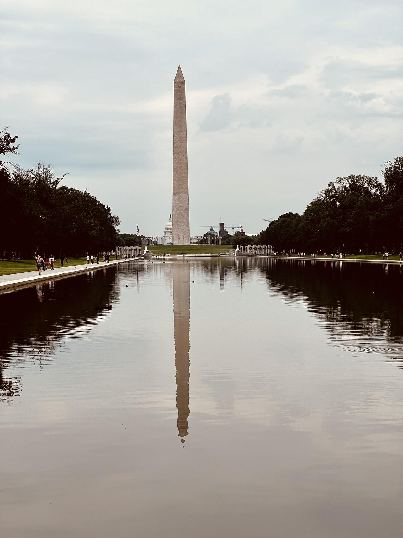 Obelisk reflecting in long pool, green trees, overcast sky, distant people.