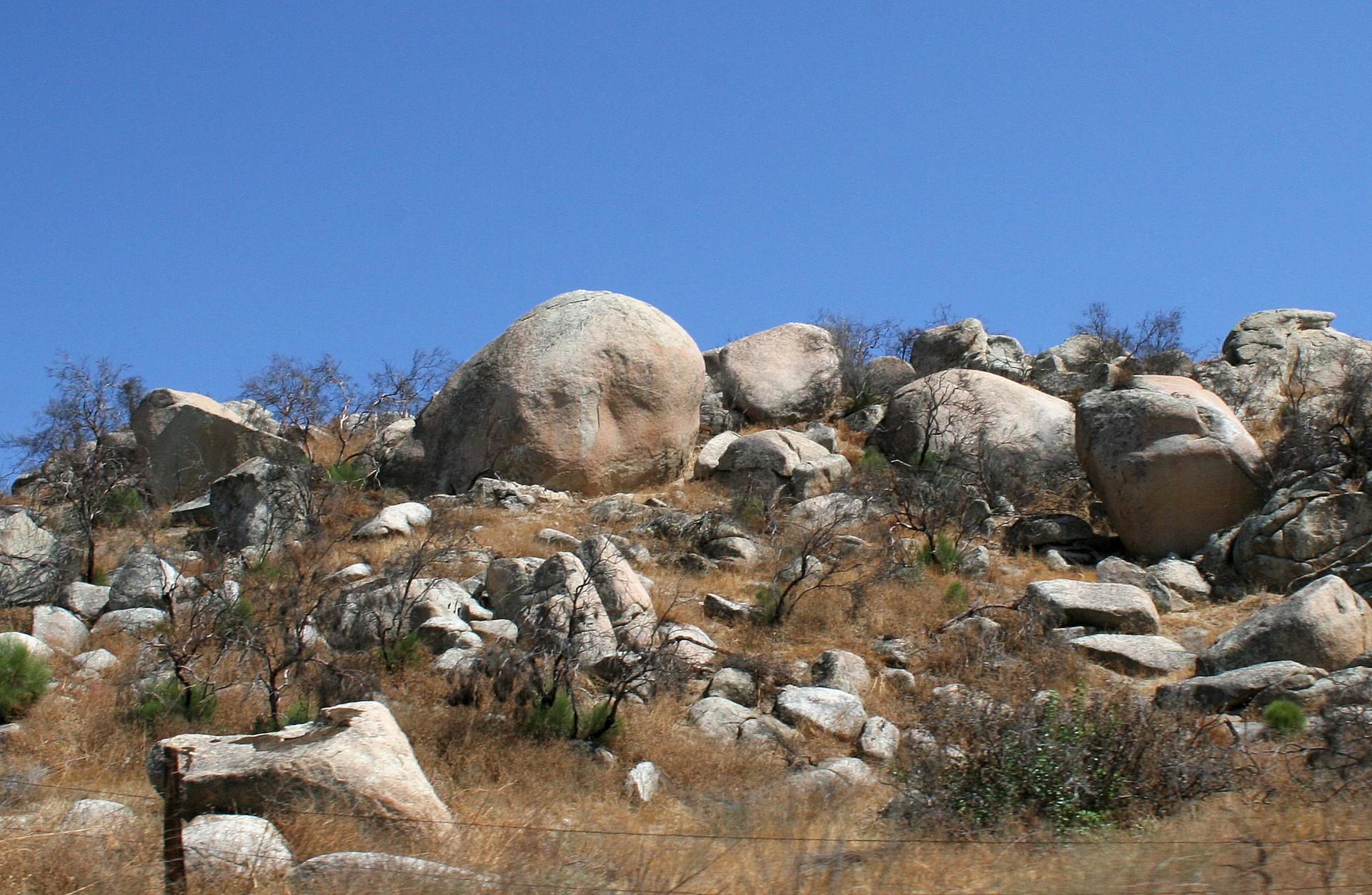 Sunny rocky hillside with large boulders, dry grass, and sparse trees under blue sky.