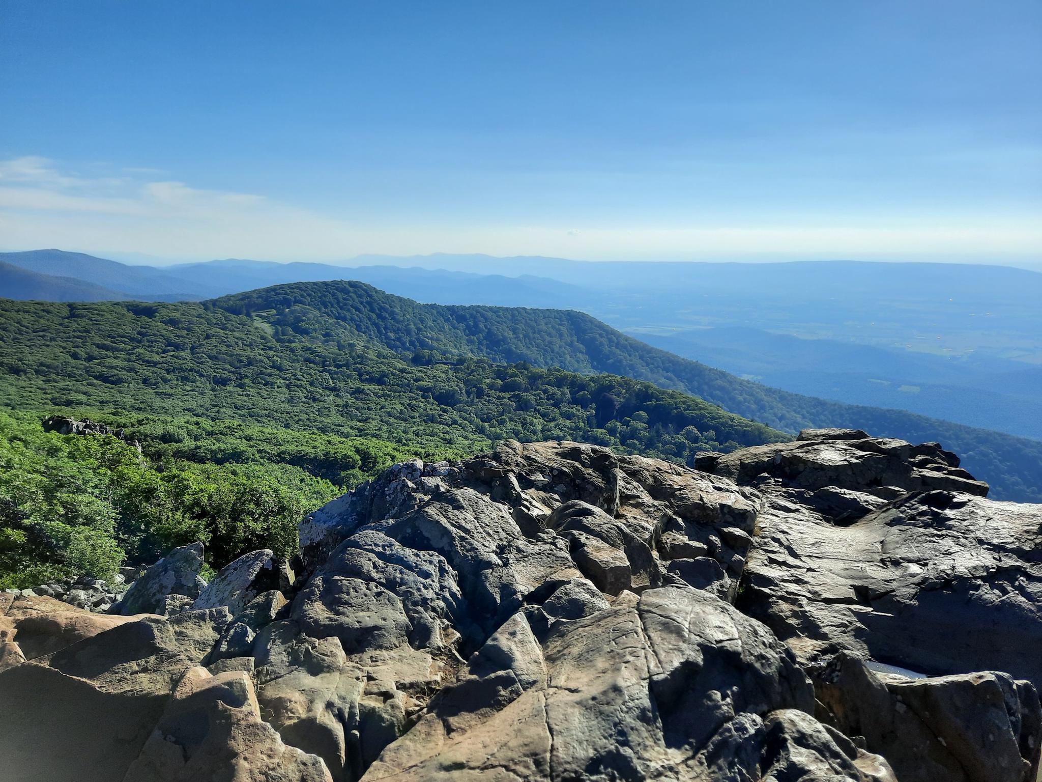 Rocky foreground with green mountains and hazy distant landscape under clear sky.