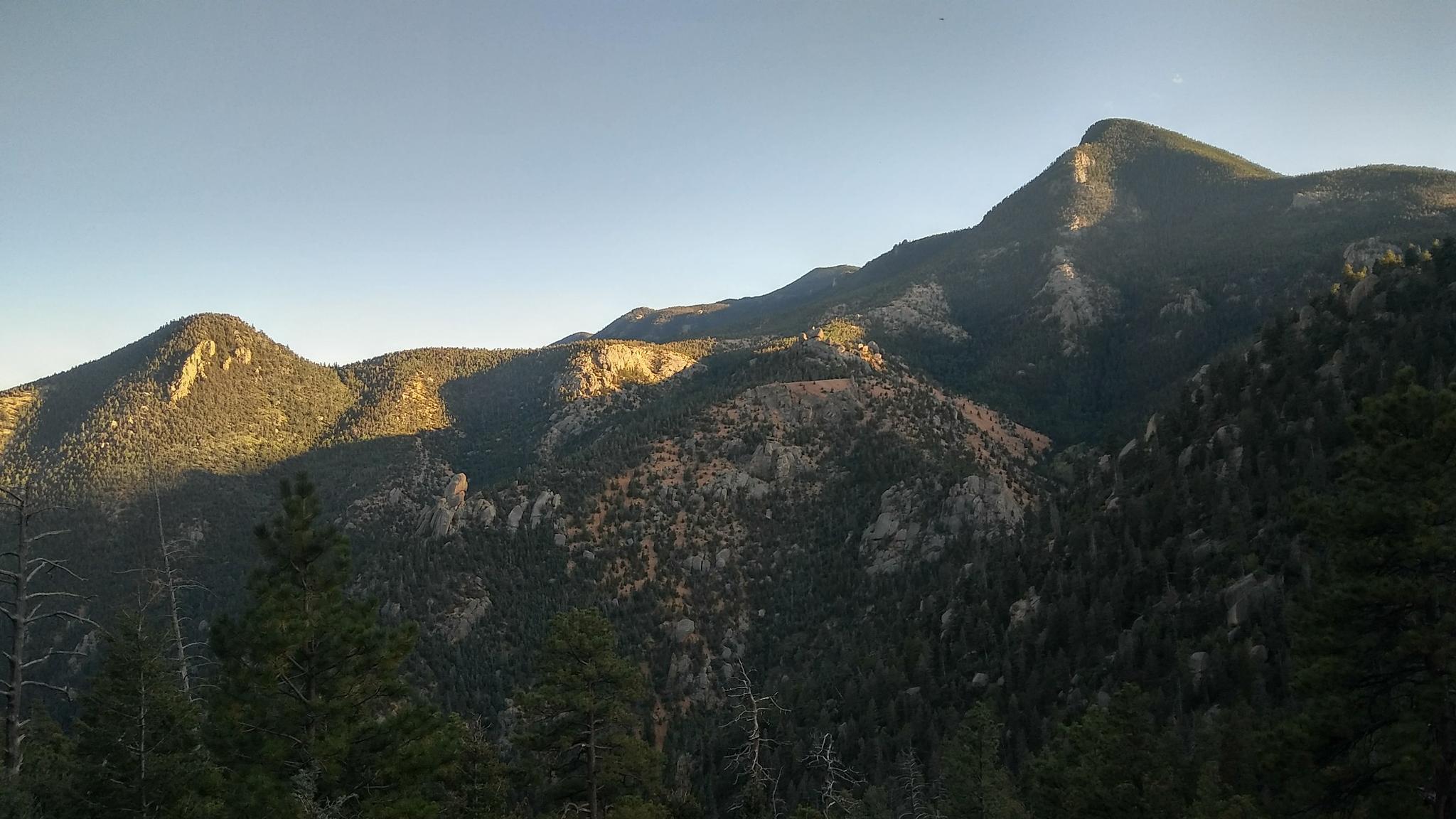 Golden light illuminates forested mountain peaks under a clear evening sky.