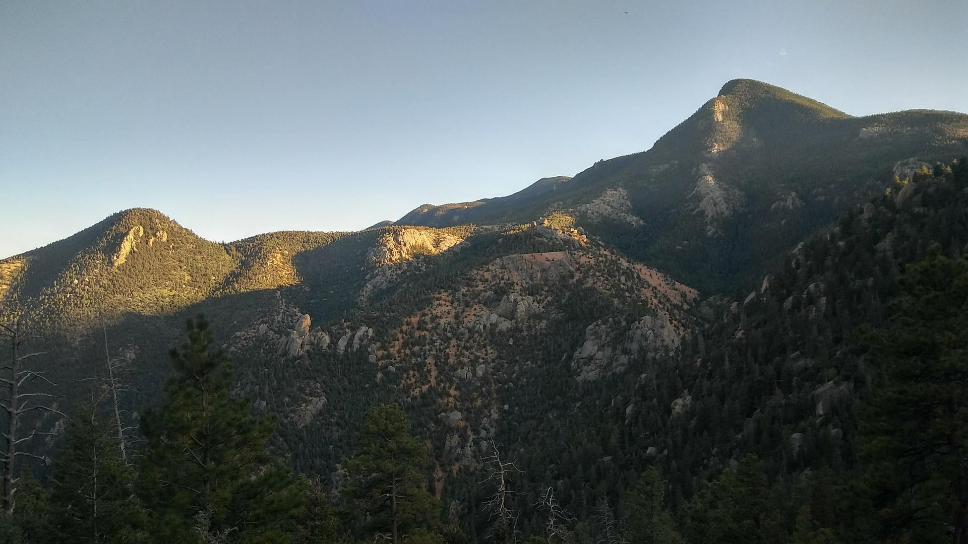 Golden light illuminates forested mountain peaks under a clear evening sky.