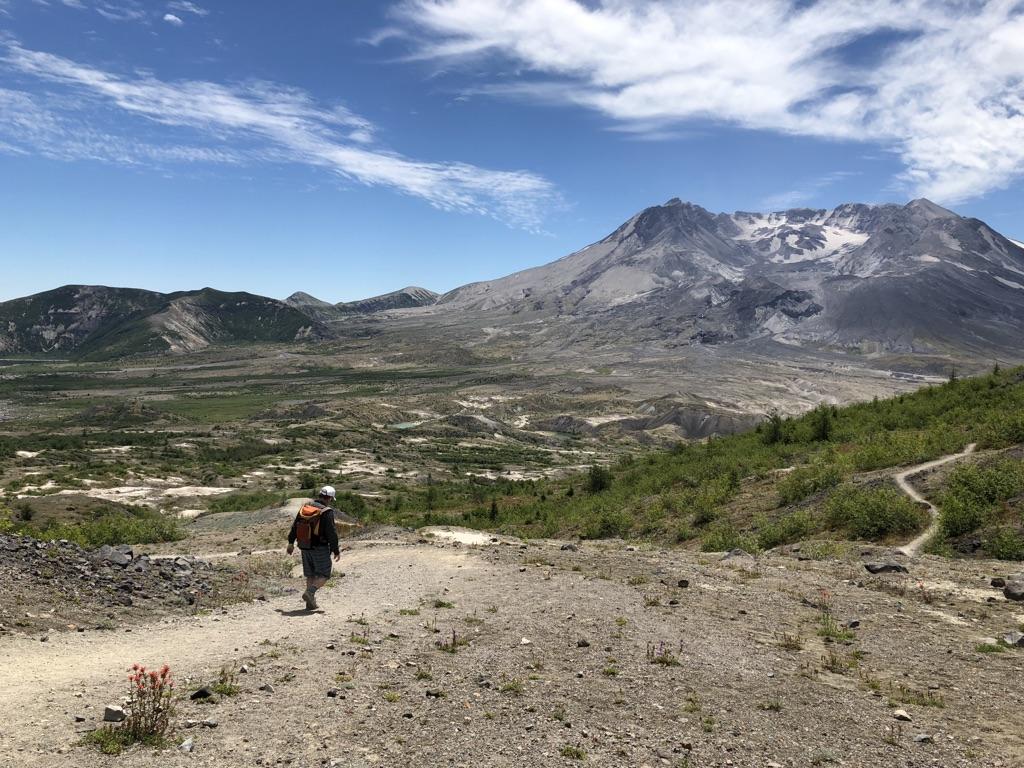 A hiker walks a dirt trail towards a large, grey mountain on a sunny day.
