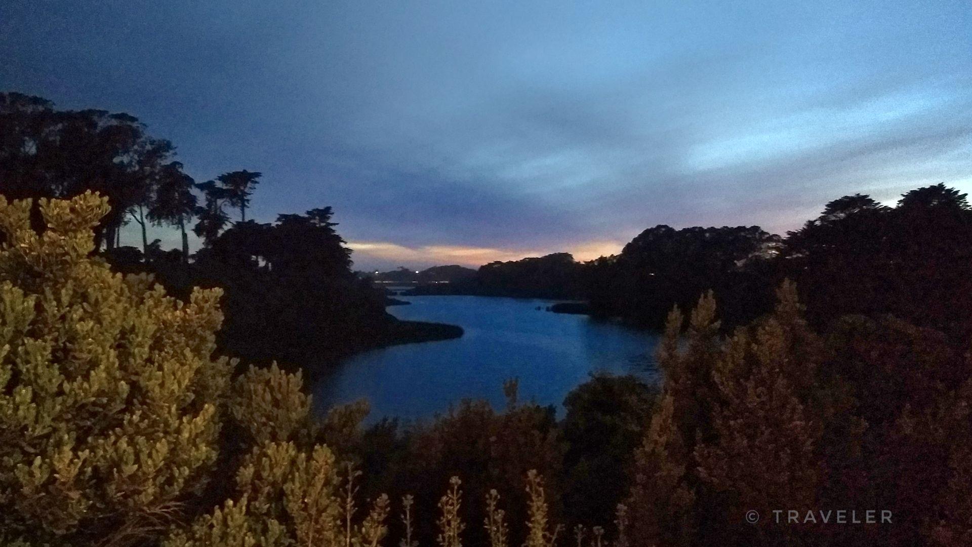 A dark, serene lake surrounded by silhouetted trees at dusk with a colorful sky.