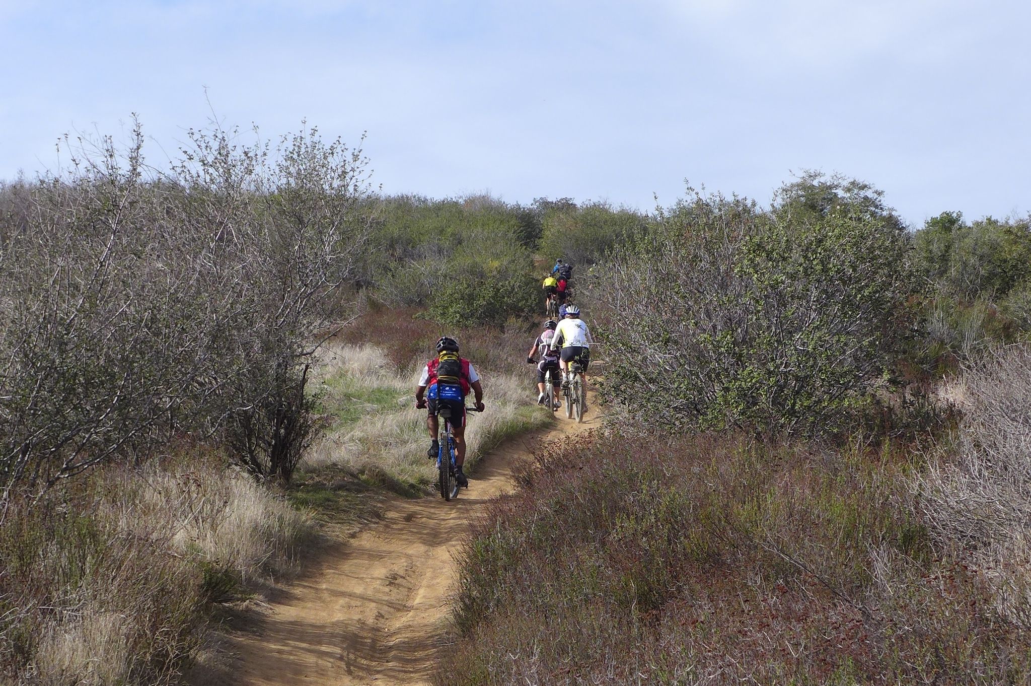 Group of mountain bikers riding a dirt trail through dry, bushy hills. - Crafton Hills Loop