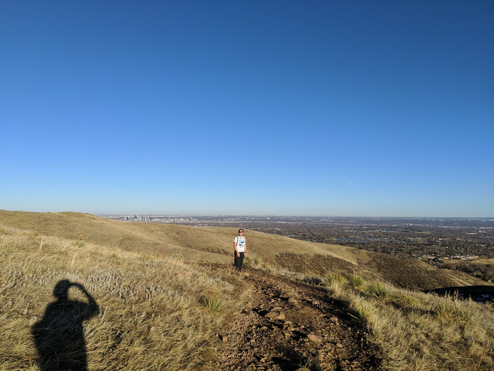 Person hiking on a sunny, dry hillside with a distant city view. - Green Mountain and Hayden Trail Loop