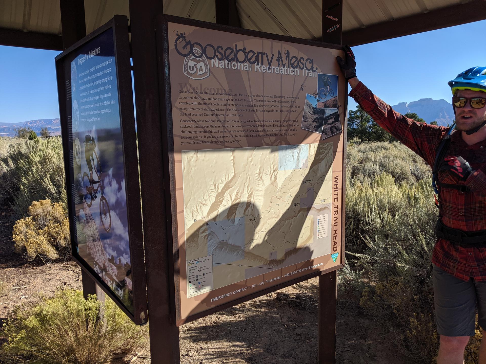 Man in helmet and sunglasses next to a large trail information sign.