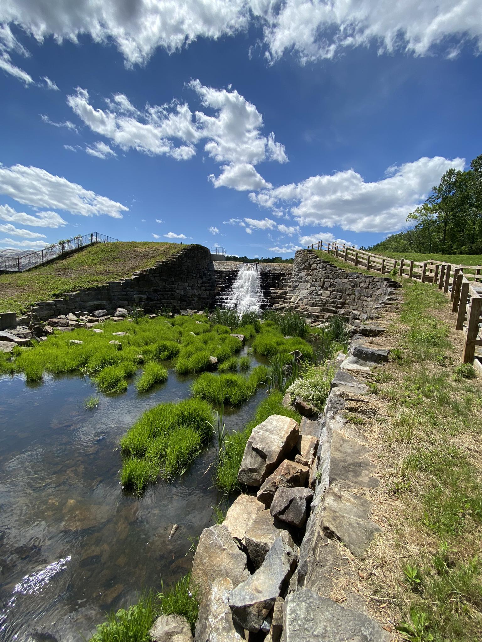 Waterfall cascading over stone steps into a grassy stream under a bright sky.