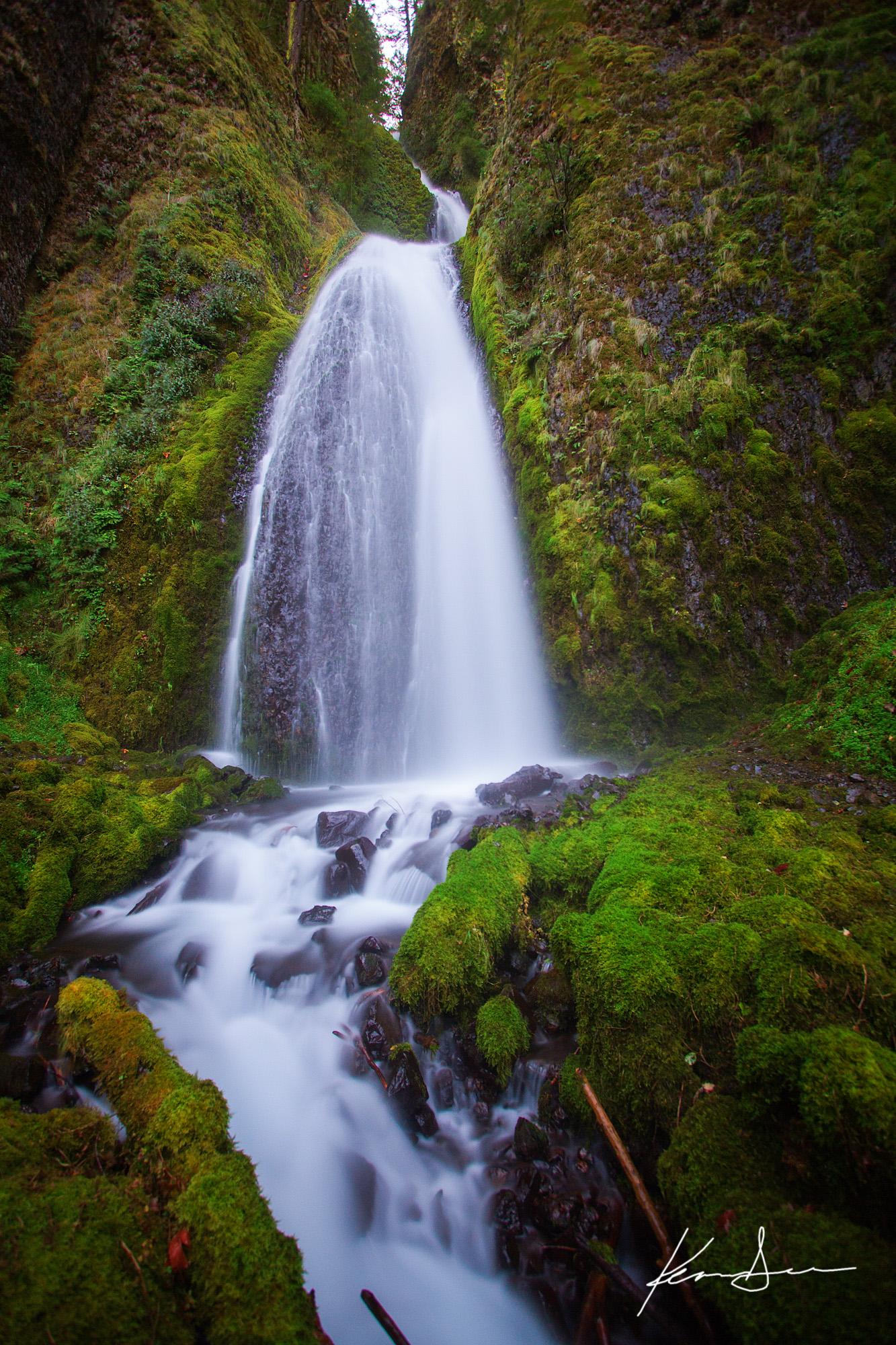 A tall waterfall cascades over mossy rocks into a stream in soft light.