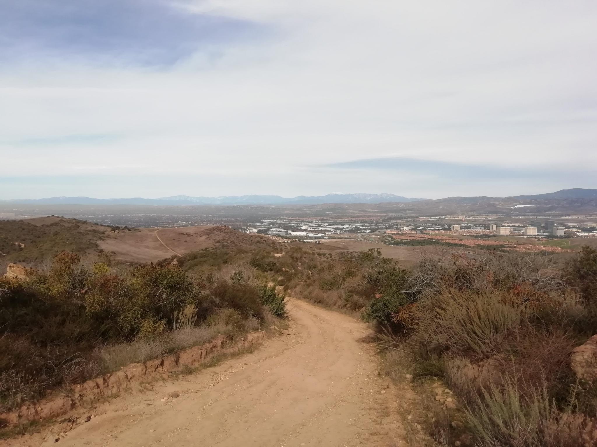 Wide dirt road descends through dry brush towards distant city under cloudy sky.