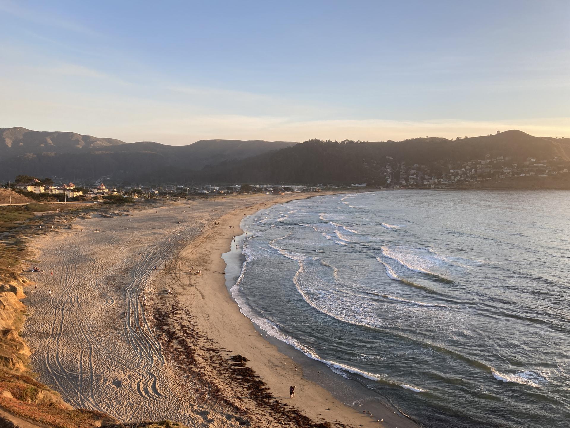 Long sandy beach with gentle waves and distant hills at sunset.