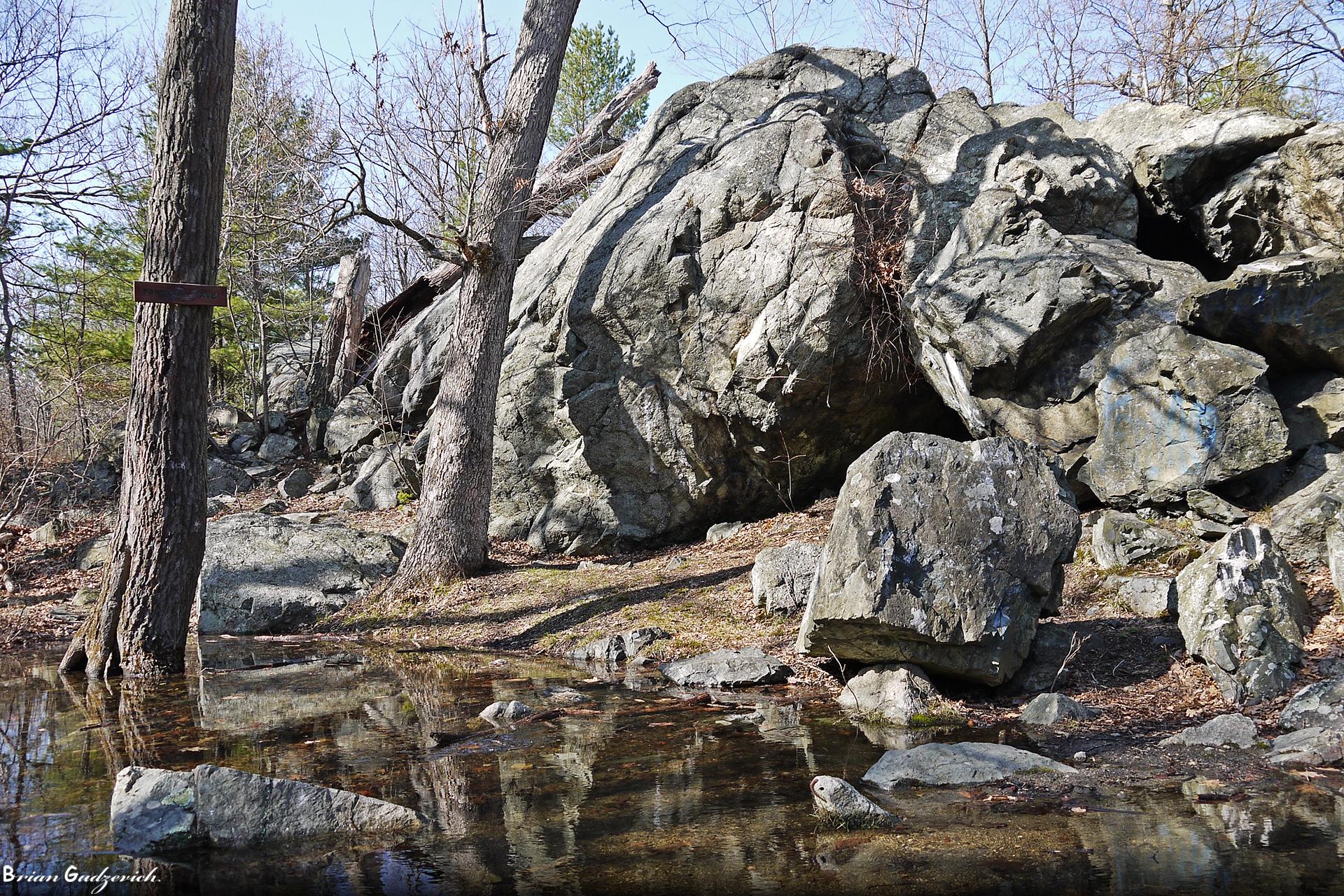 Large boulders and trees reflected in still water in a wooded area.