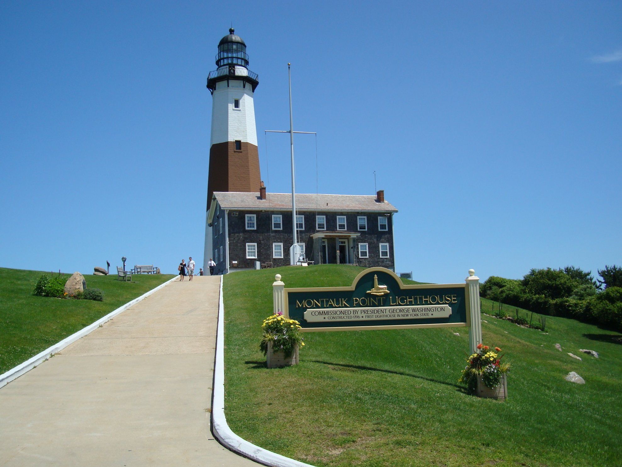 Lighthouse and building on a green hill with a sign under a clear sky. - Paumanok Path: Montauk Point Loop