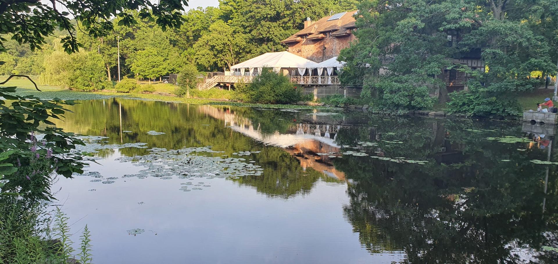 White tent and building reflected on a calm lake surrounded by green trees.
