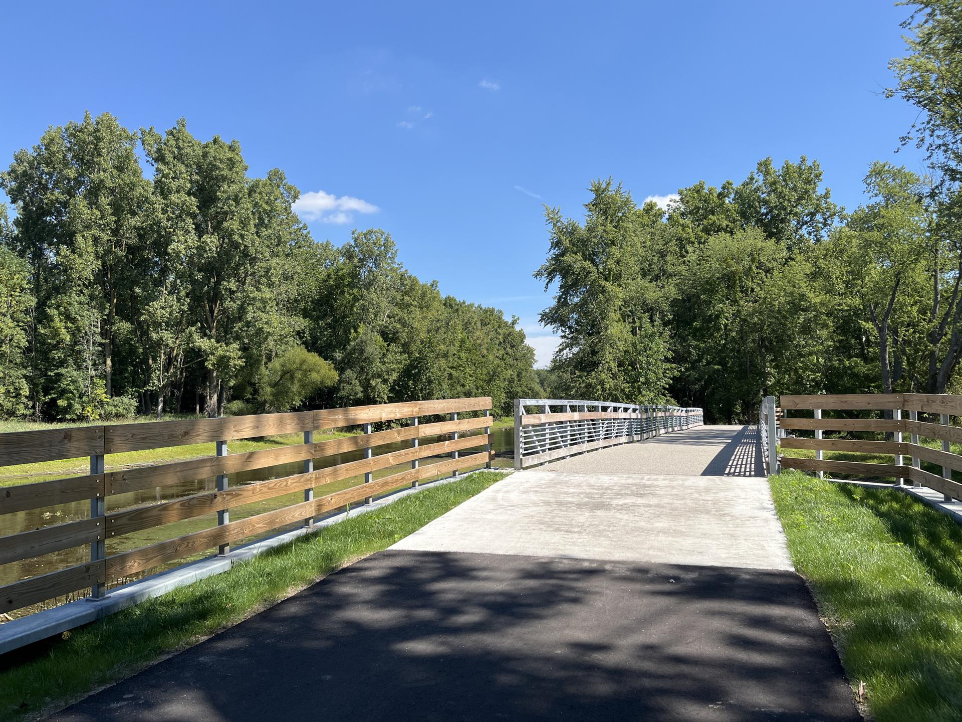 Paved path leads to a modern bridge over water, surrounded by lush green trees.