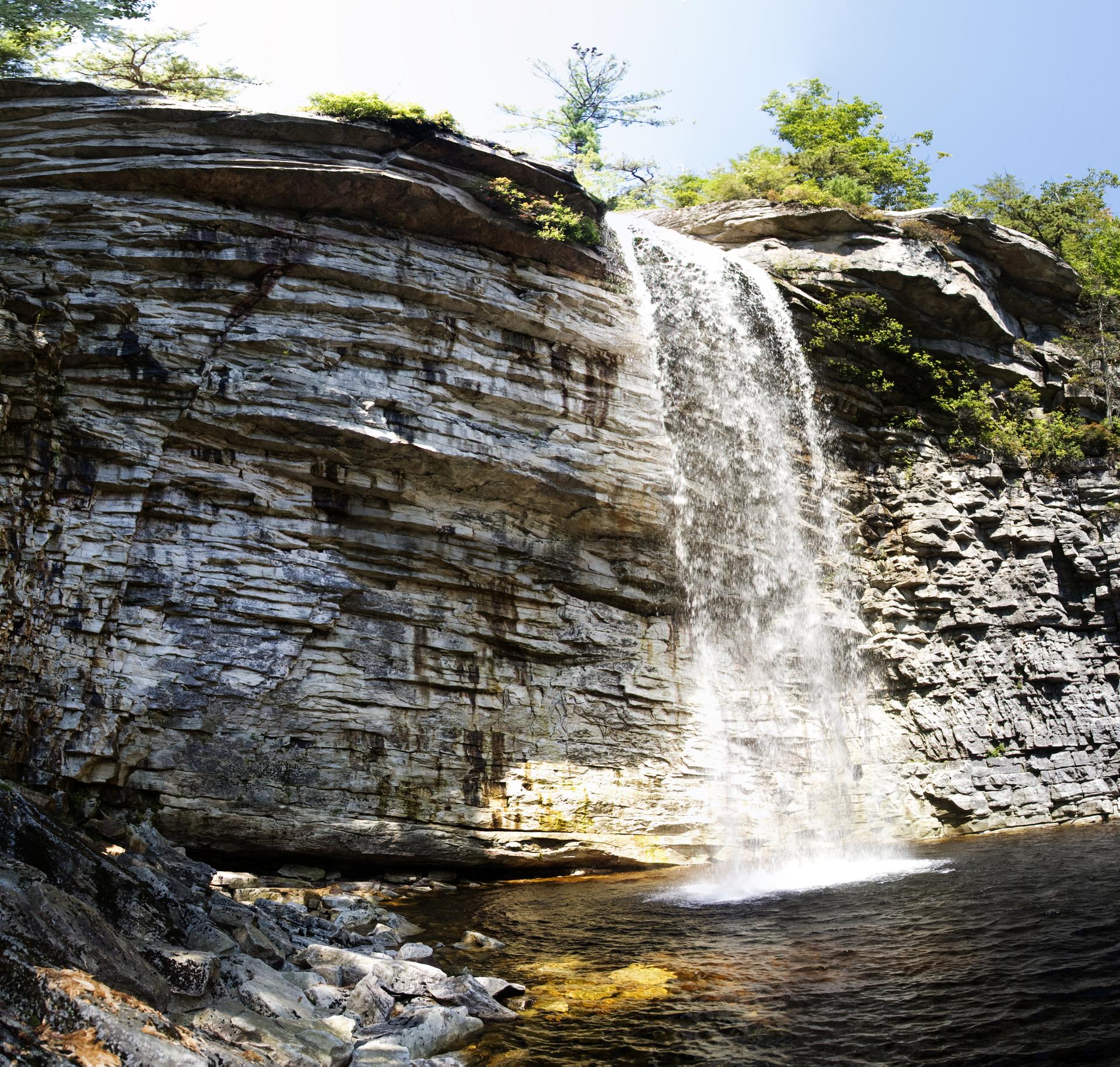 Tall waterfall cascades into a dark pool surrounded by rocky cliffs on a bright day.