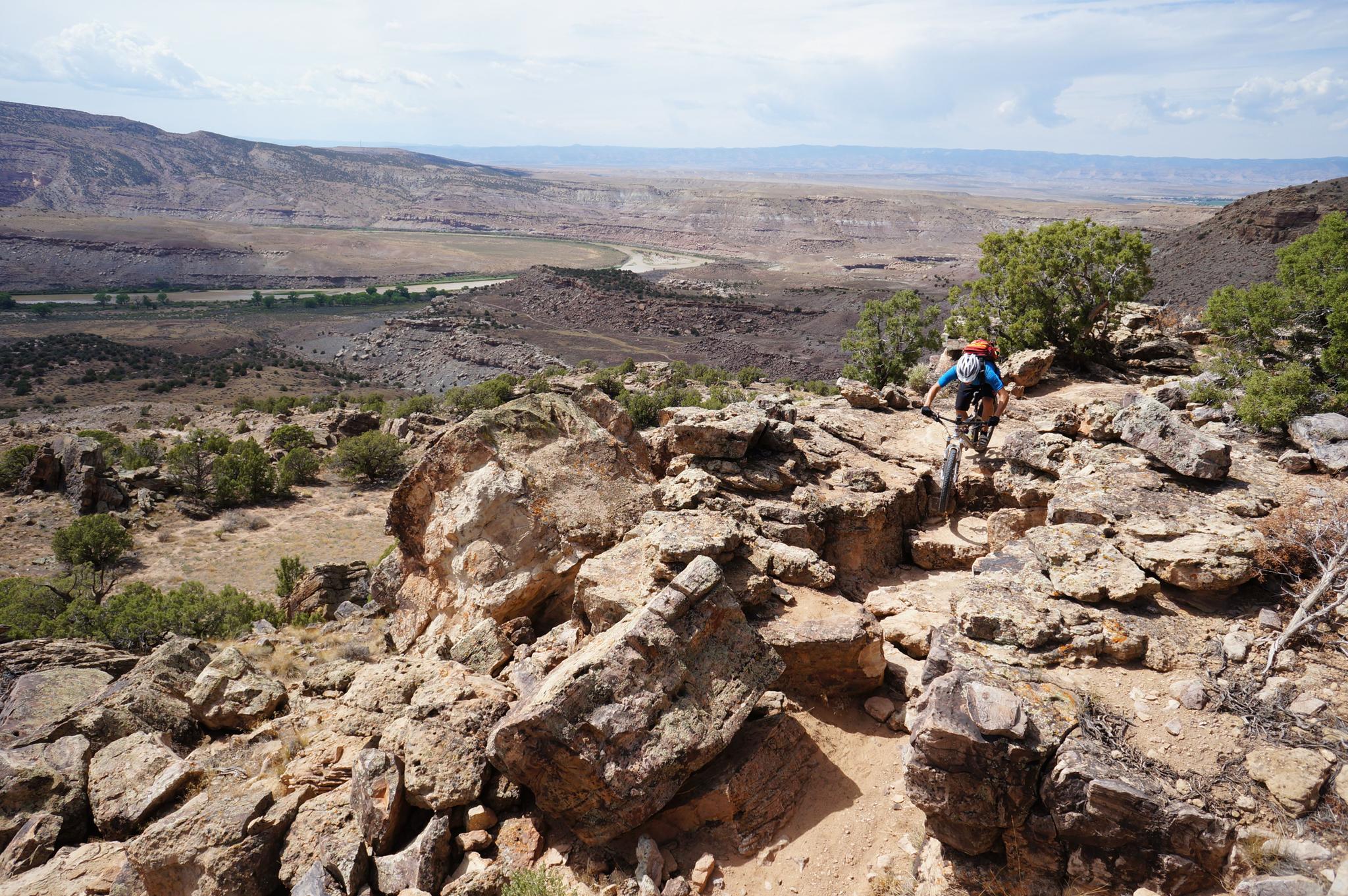 A mountain biker navigates a rocky trail overlooking a vast desert valley.