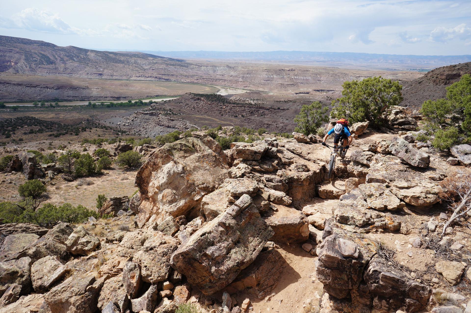 A mountain biker navigates a rocky trail overlooking a vast desert valley.