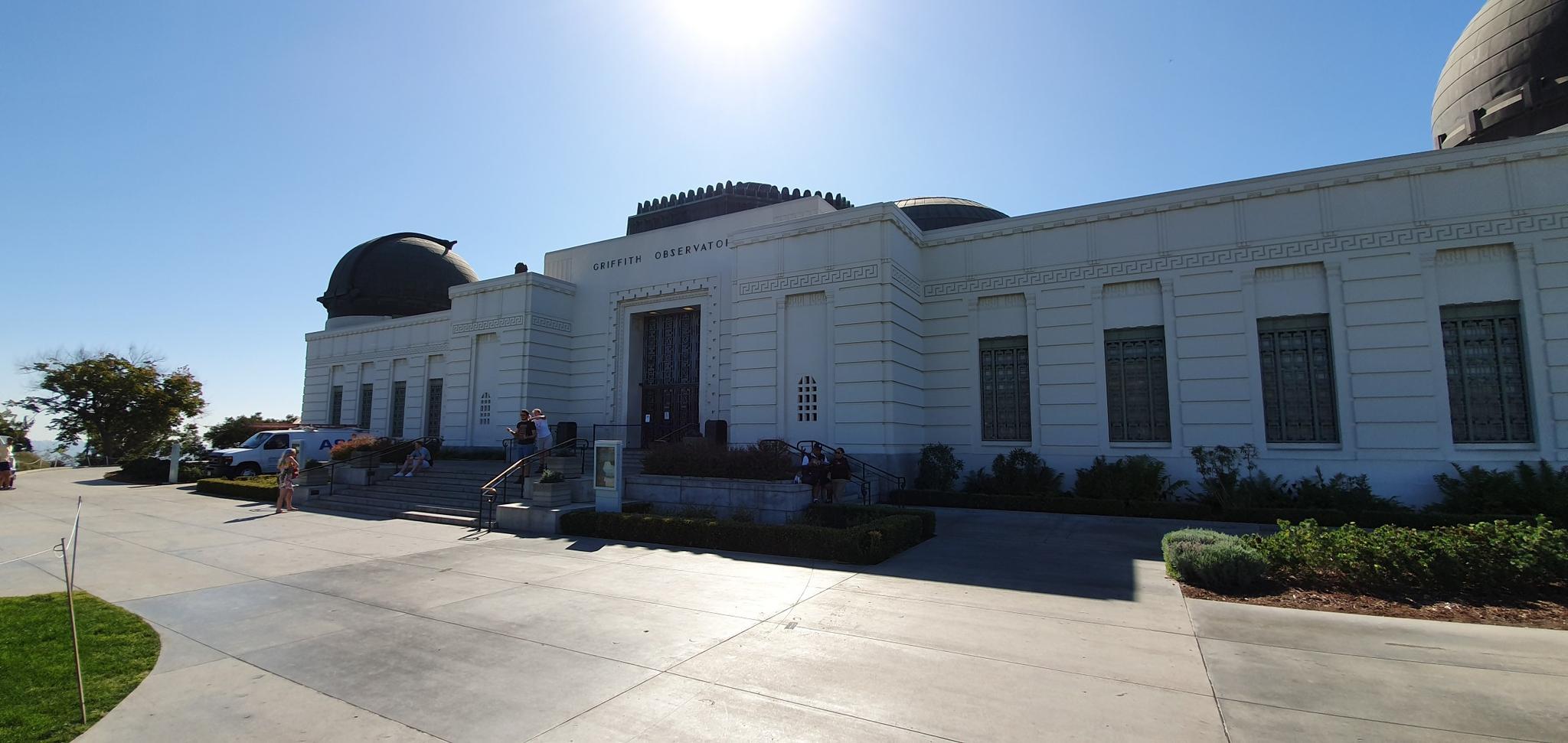 Large white building with domes under bright sun, people walking on pavement.