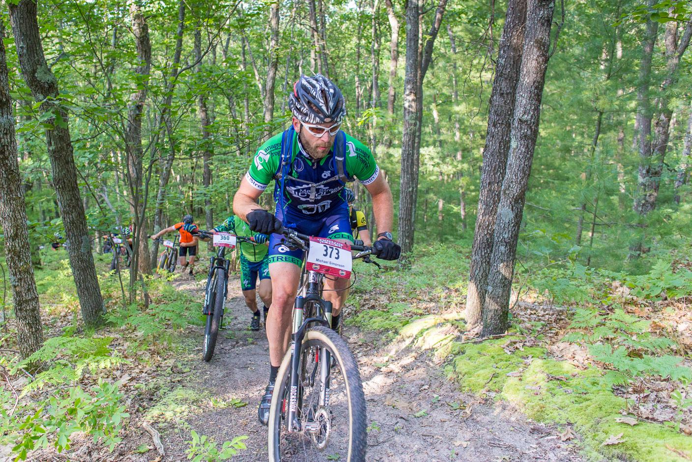 Mountain biker in green kit rides uphill on a sunlit forest path.