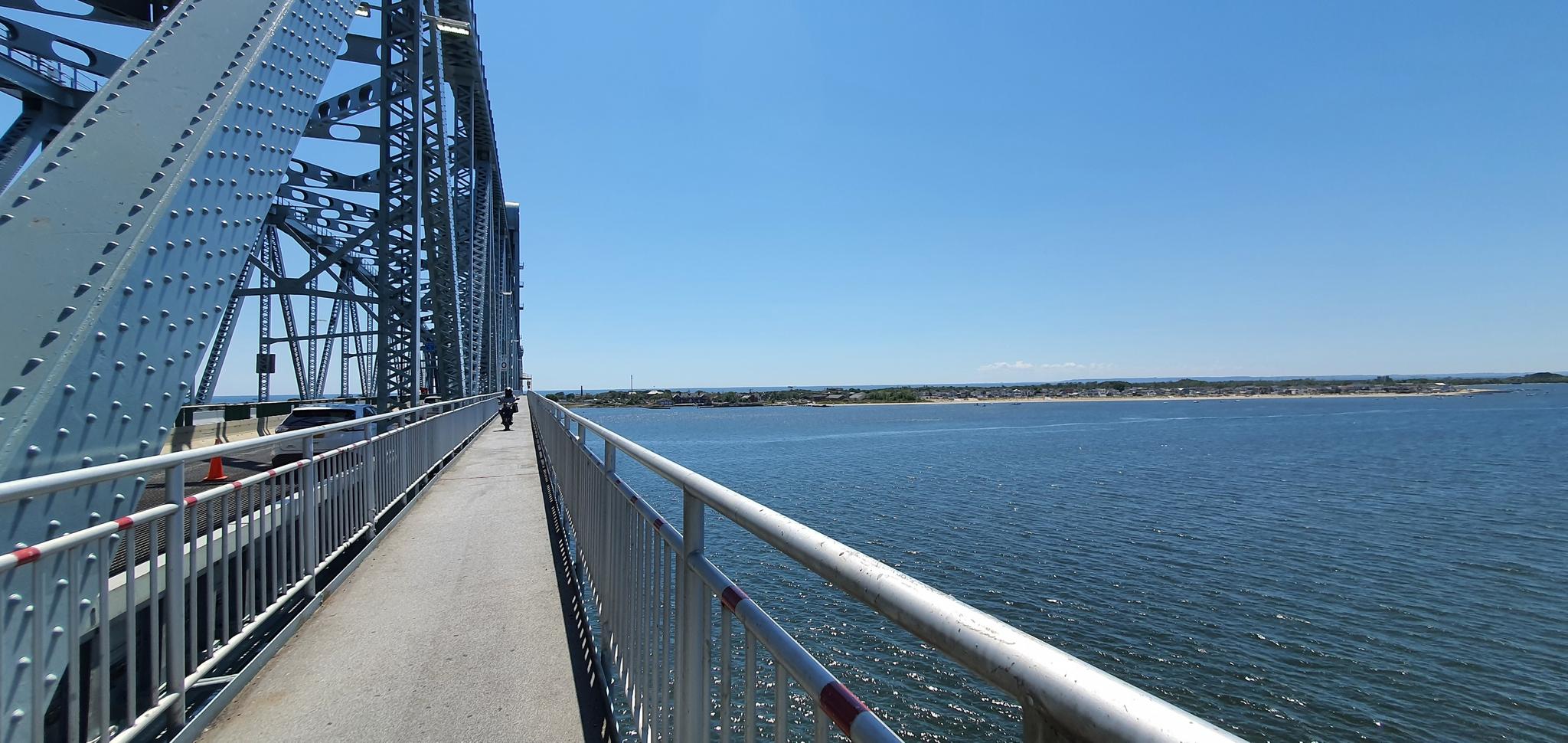A person walks on a bridge next to blue water under a clear sky.