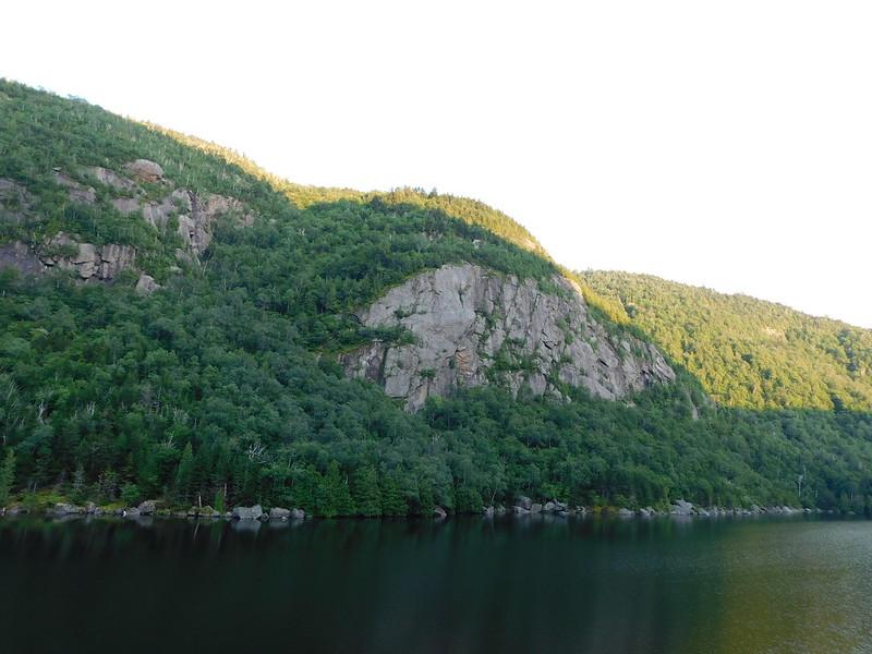 Steep, forested mountain with rocky cliffs overlooking dark water under bright sky.