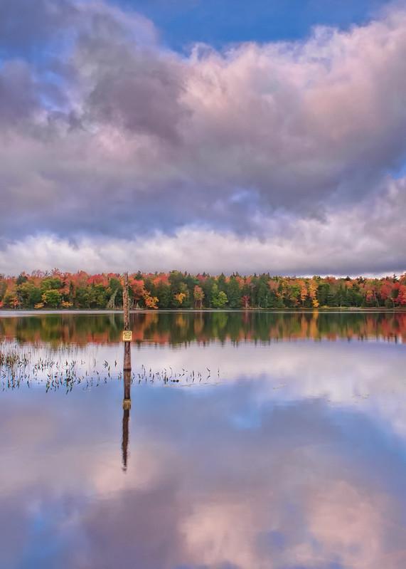 Calm lake reflecting cloudy sky and vibrant autumn trees with a wooden post.