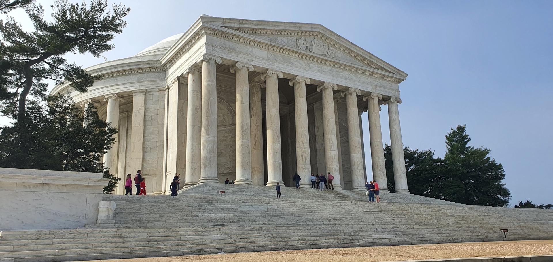 People walk on the steps of a grand, white, columned building on a sunny day.