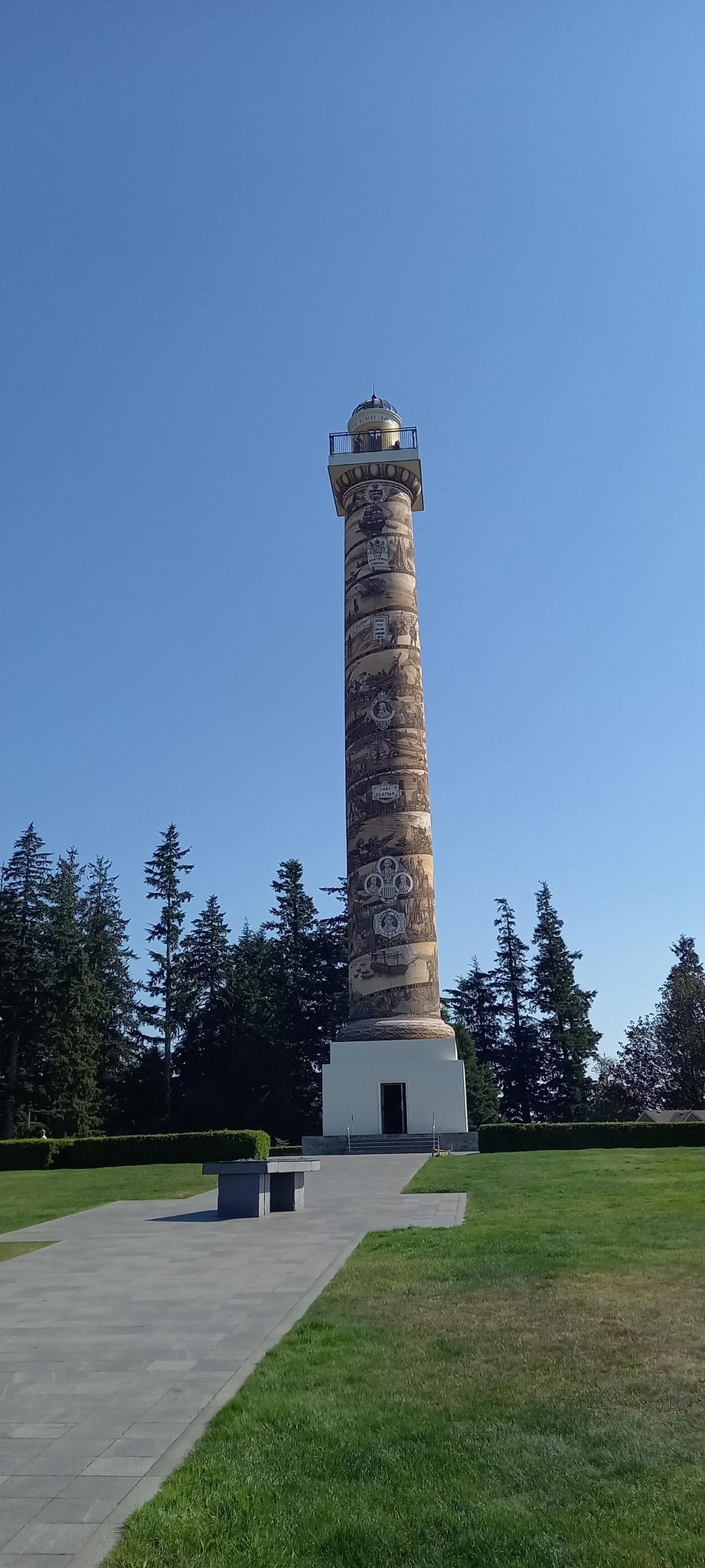 A tall, decorated column monument stands on a green lawn under a clear sky.