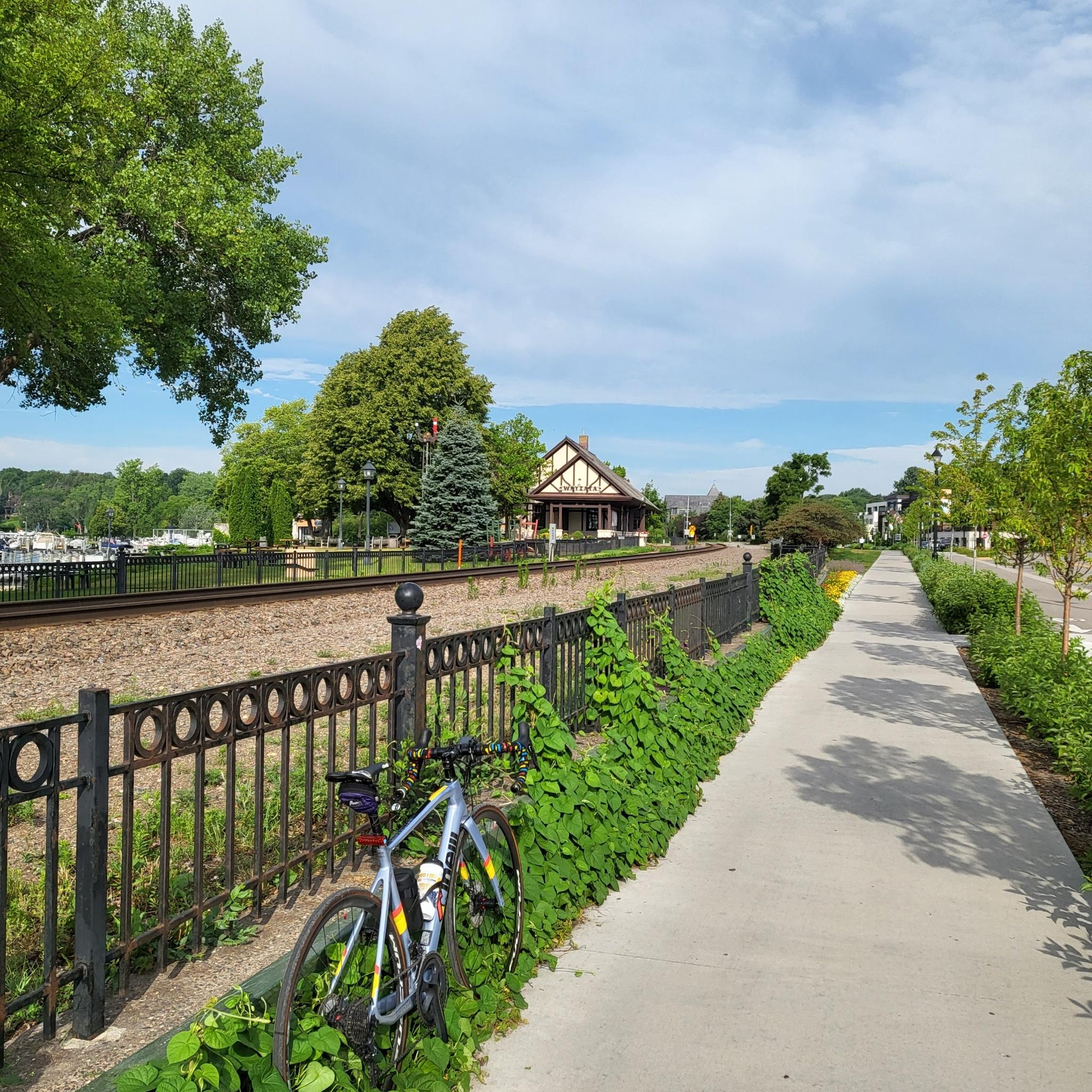 Bicycle parked beside a paved path next to railroad tracks on a sunny day.