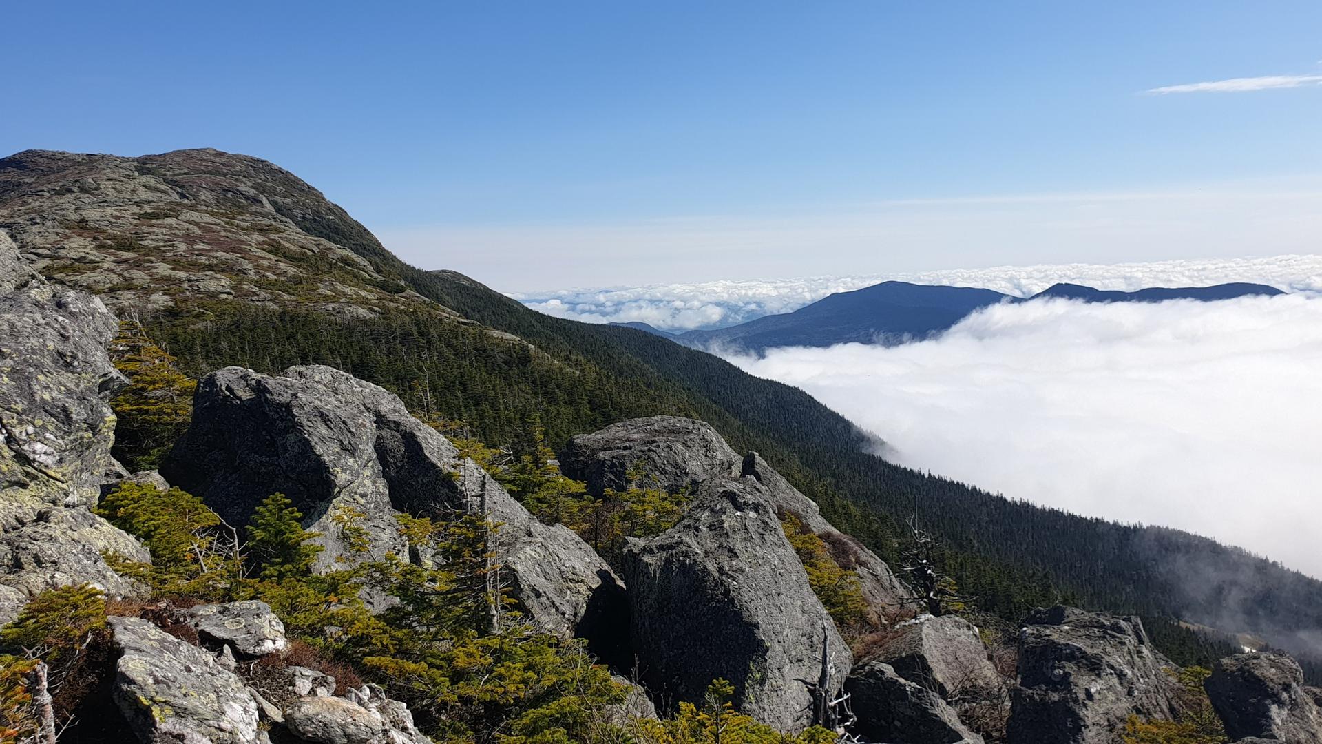 Sunny mountain ridge with large rocks, green trees, and clouds below.