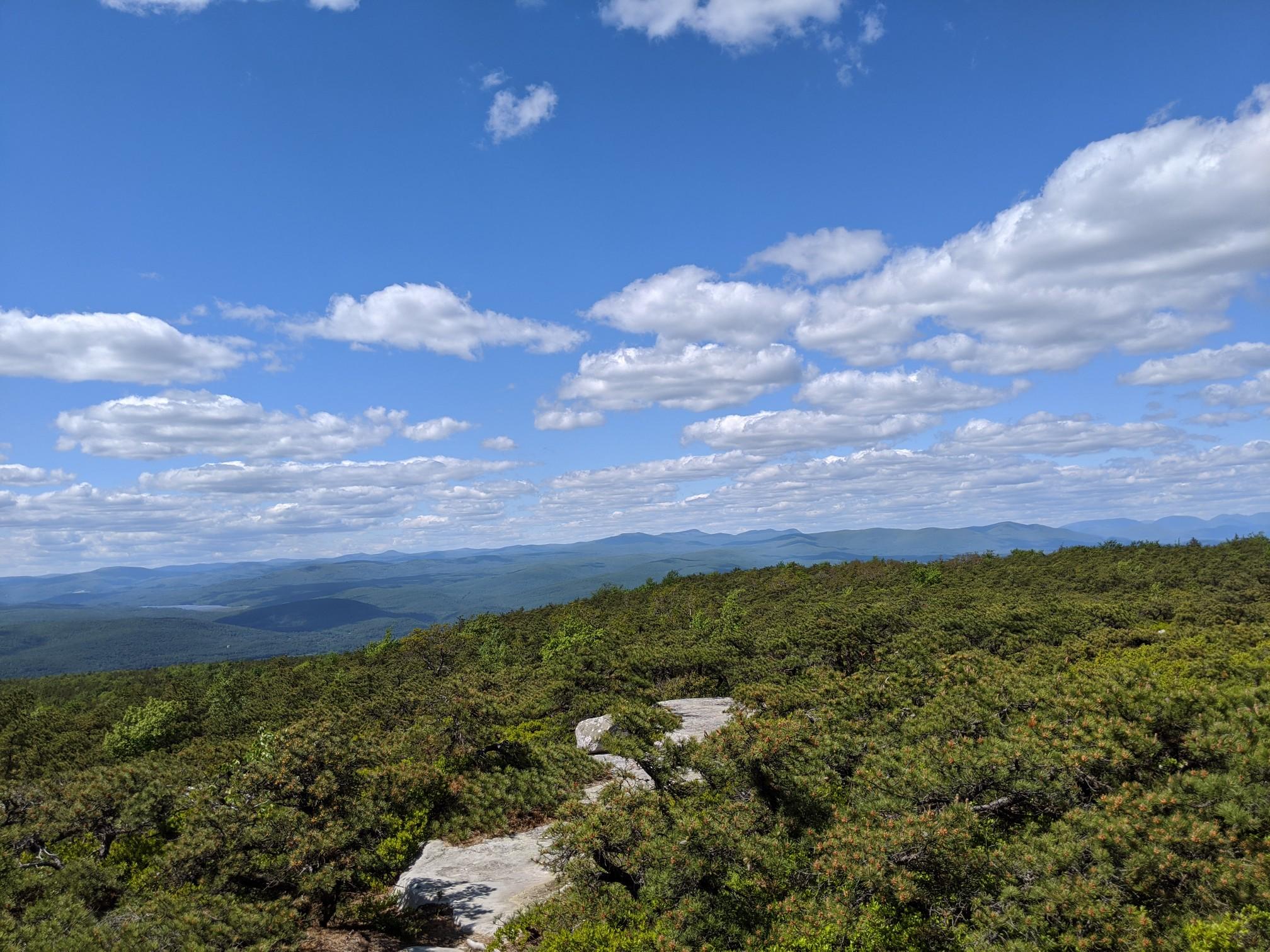 Expansive view of green forested hills and distant mountains under a cloudy blue sky.