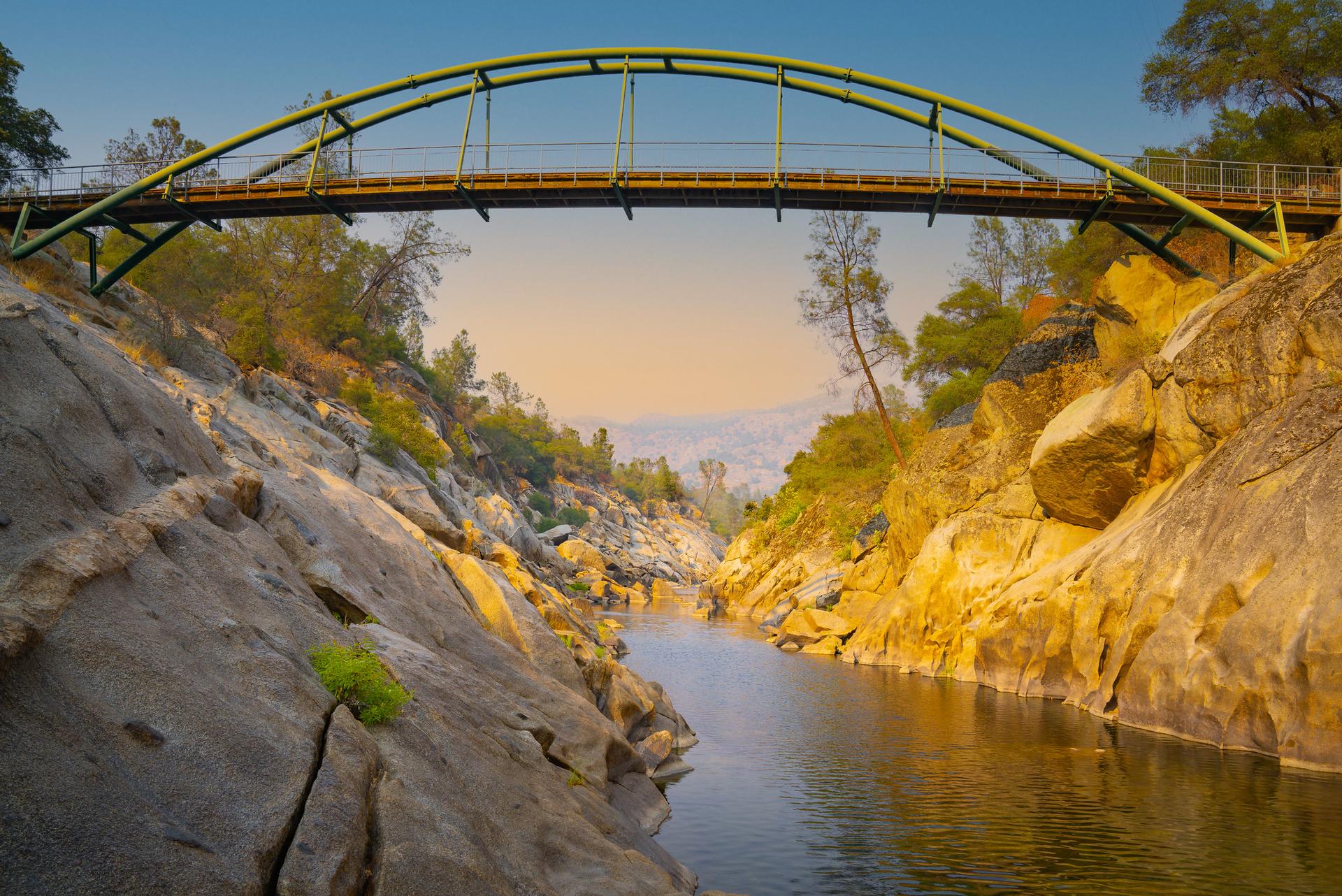 Green arched bridge over a calm river in a rocky canyon at sunset.