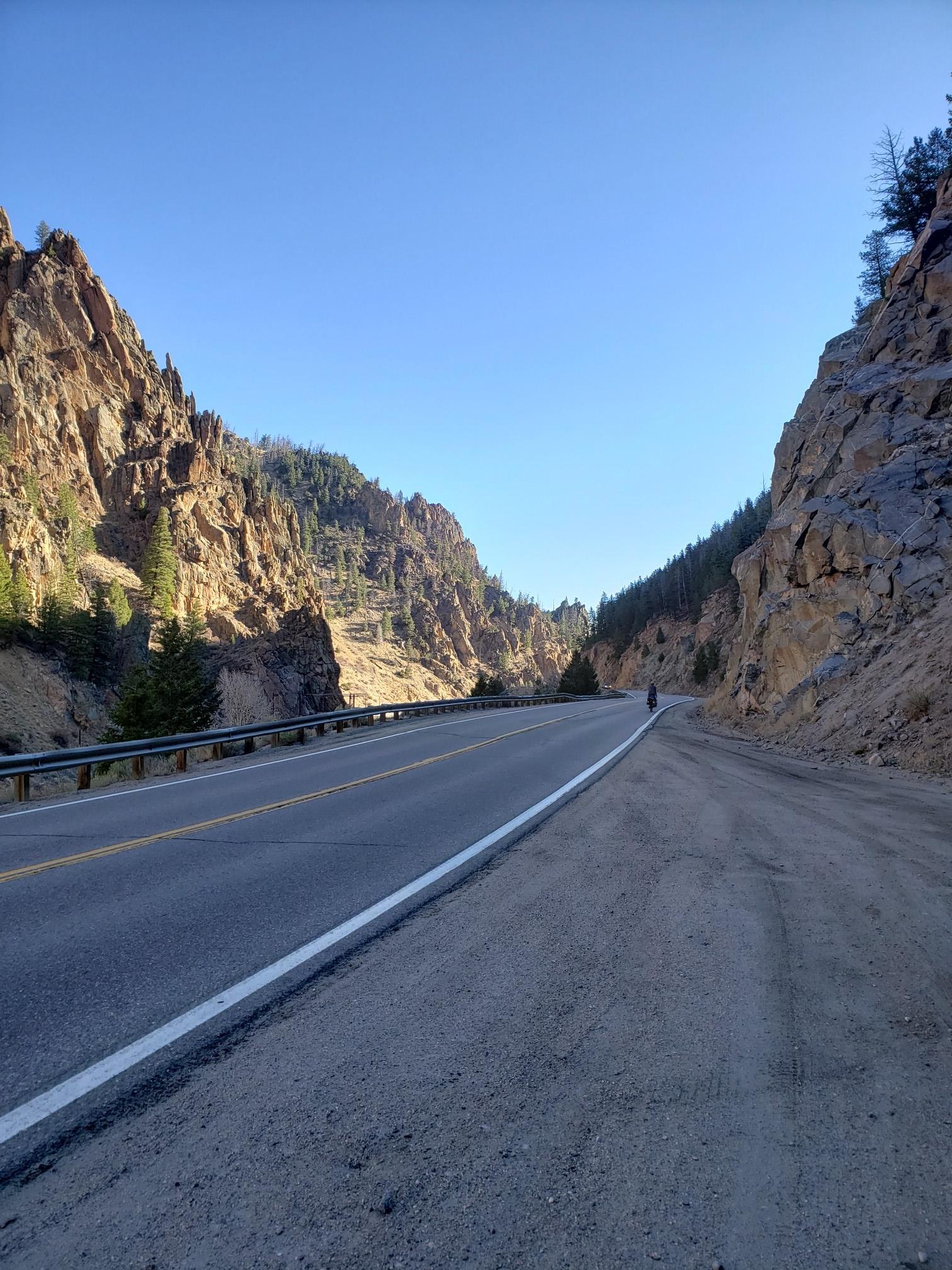 Paved road through a sunny canyon with steep rocky walls and a distant cyclist.