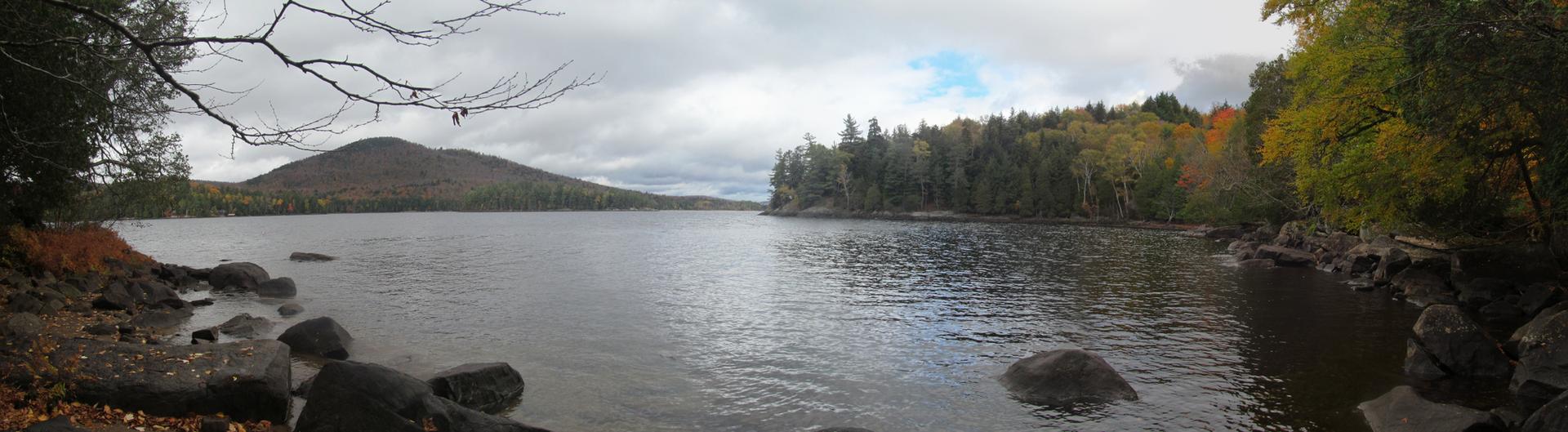 Lake with rocky shore, autumn trees, and distant mountain under an overcast sky.