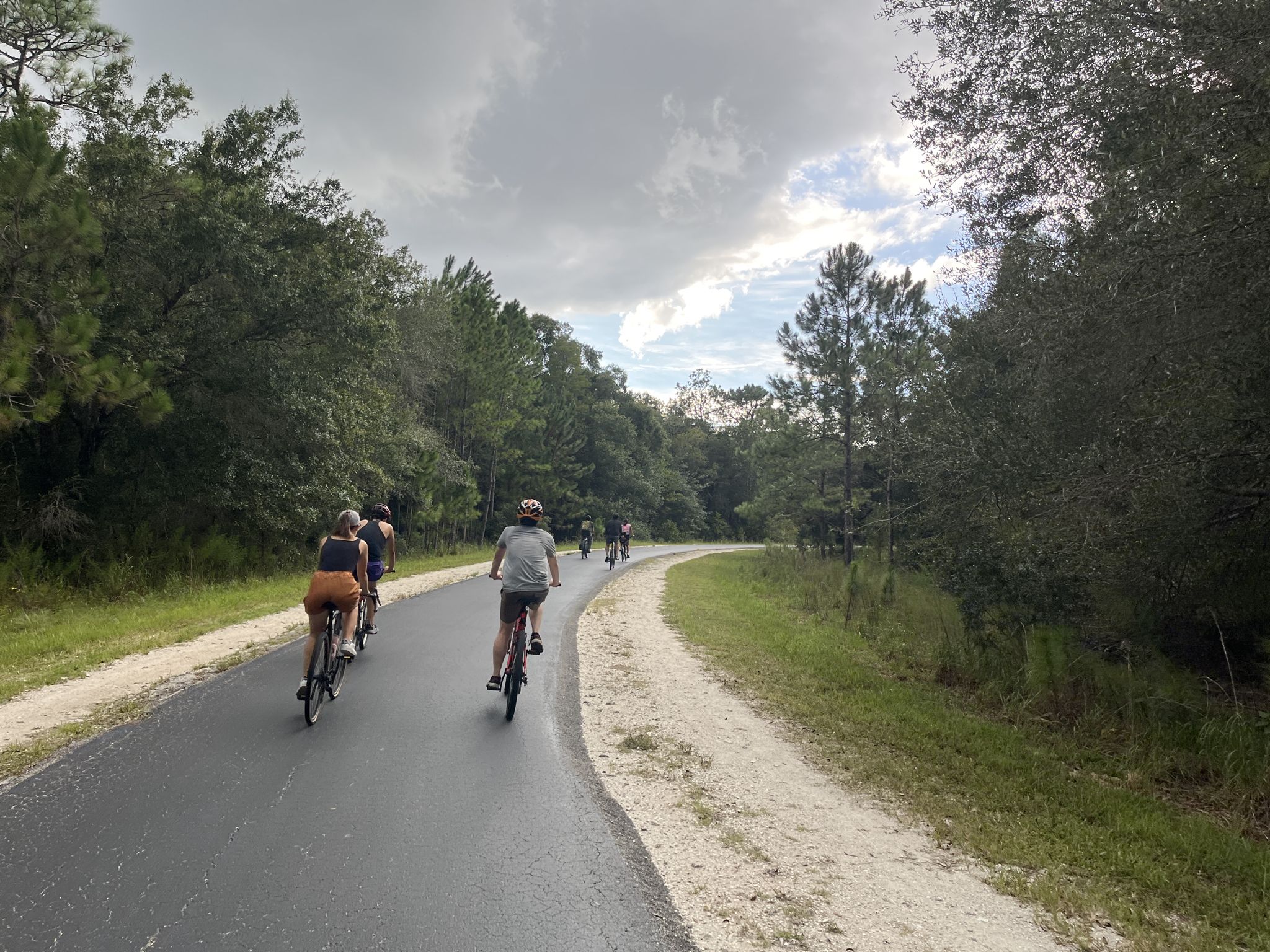 People cycling on a paved path through dense green trees under an overcast sky. - Flatwoods Park Trail Loop