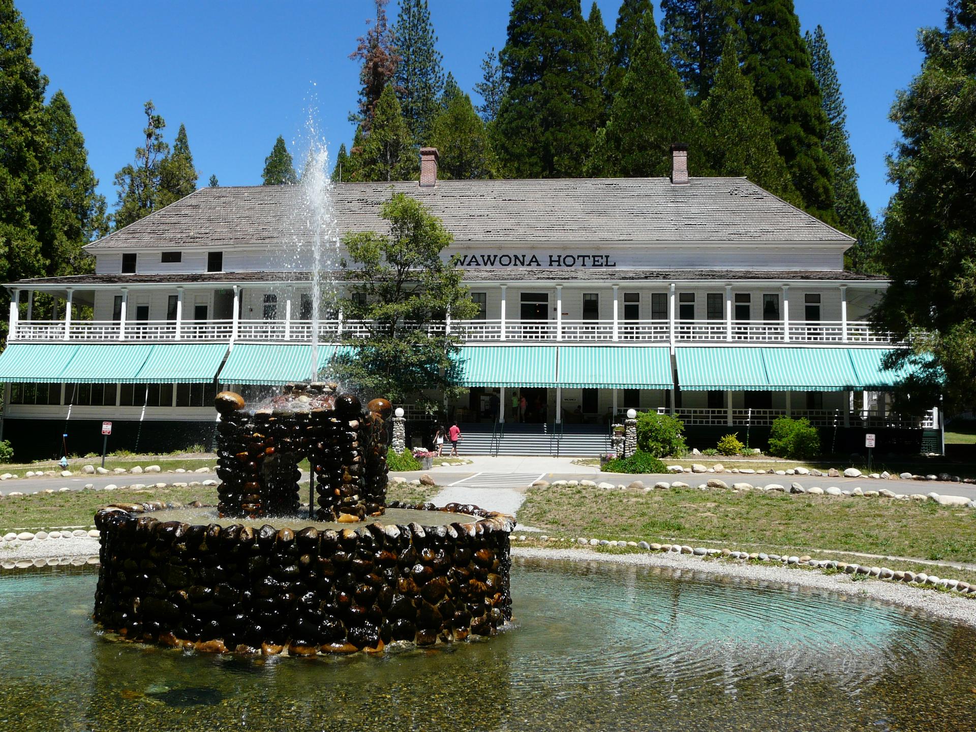 Large white hotel with green awnings, stone fountain in foreground, sunny day.
