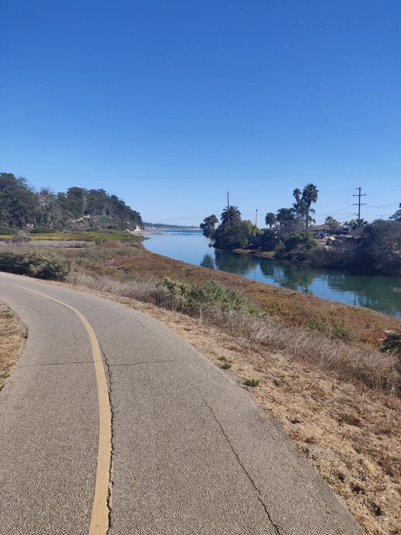 Paved path next to a calm river with lush and dry vegetation under clear sky.