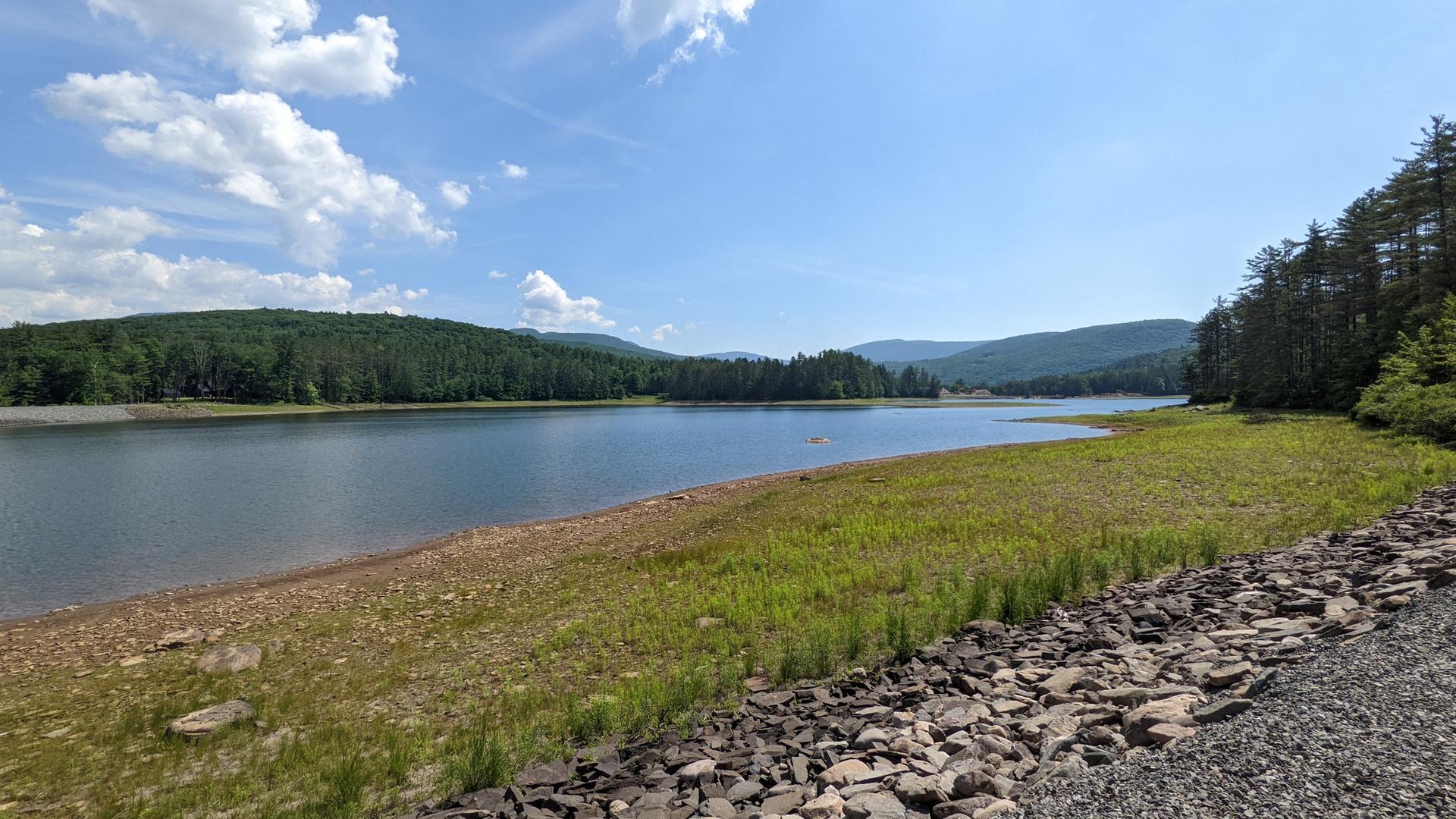 Sunny day over a wide lake with forested hills and a rocky shore.