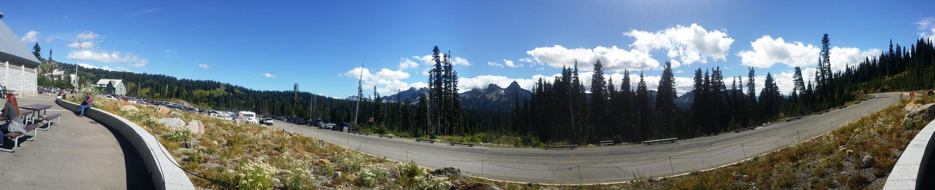 Panoramic view of a mountain road, buildings, and evergreen trees under a blue sky.